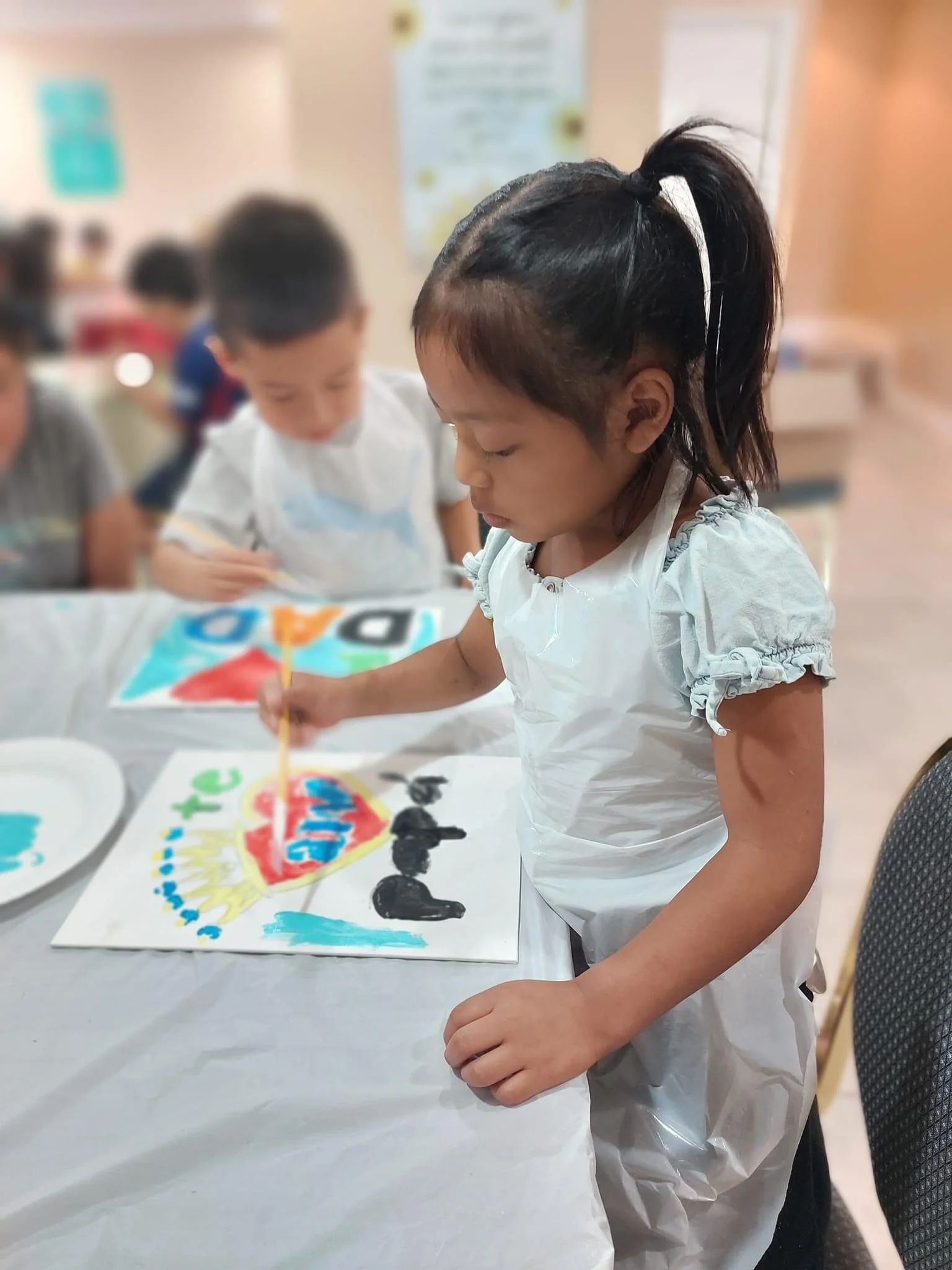 A young girl with a ponytail painting a colorful poster on a table at an indoor gathering.