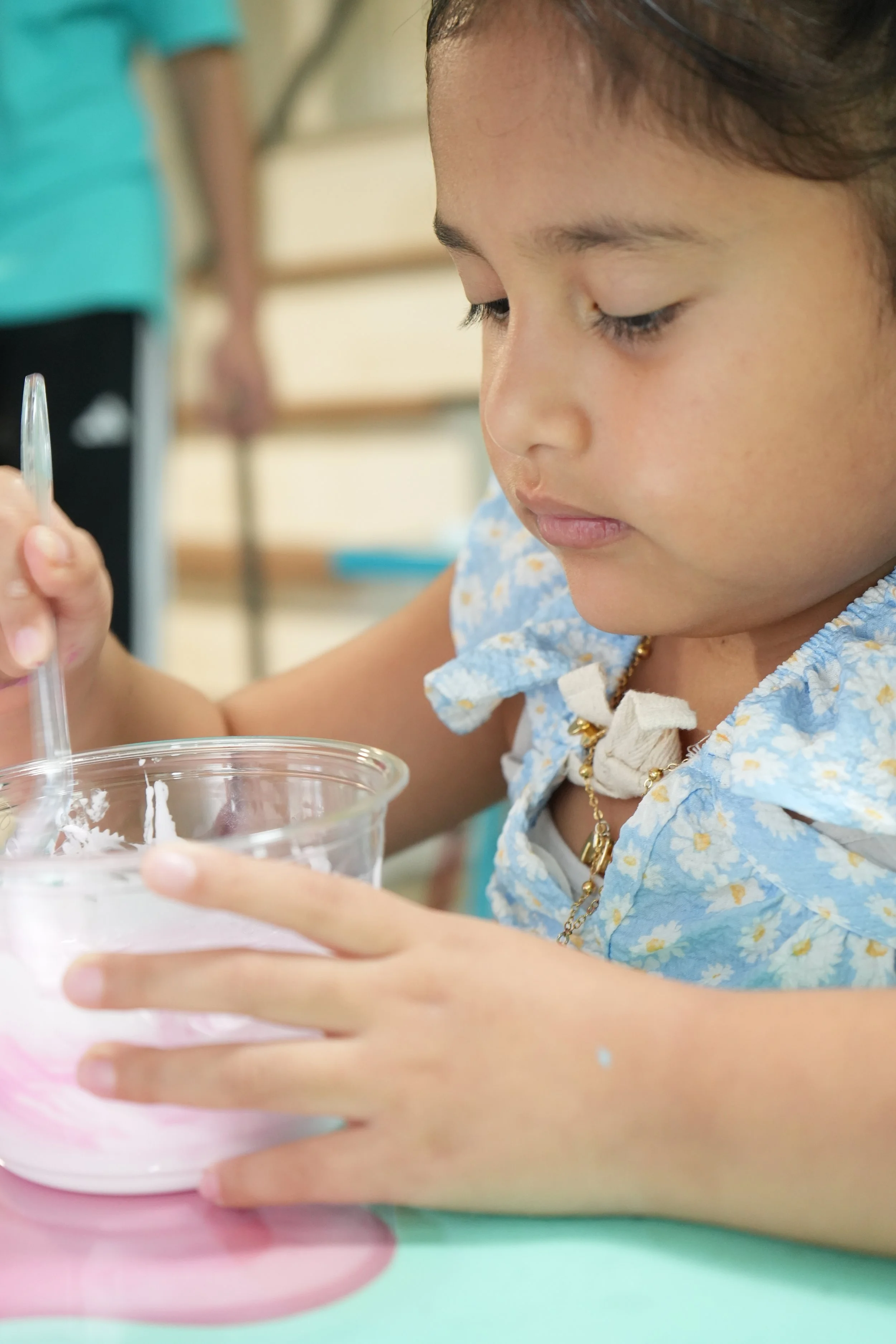 A young girl with dark hair wearing a light blue floral dress and a gold necklace, looking down and holding a plastic cup with a lid and a straw. She is focused on something inside the cup, with a blurred background.