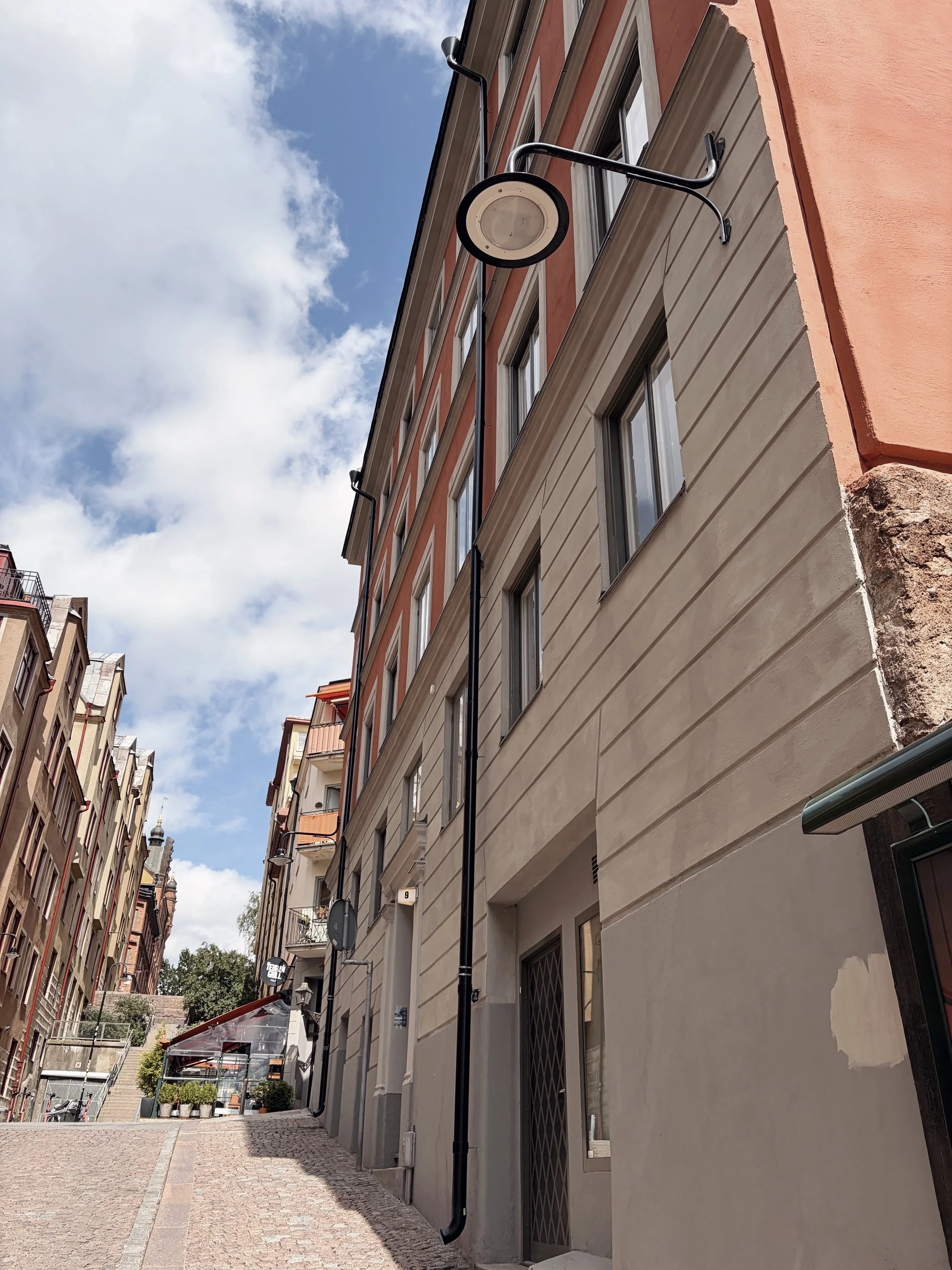 En smal gatufasad av en byggnad med flera fönster och ett rör eller ledning som hänger ner från taket. Gatans beläggning är av sten eller cobblestone. I bakgrunden syns fler byggnader och träd under en molnig himmel.
