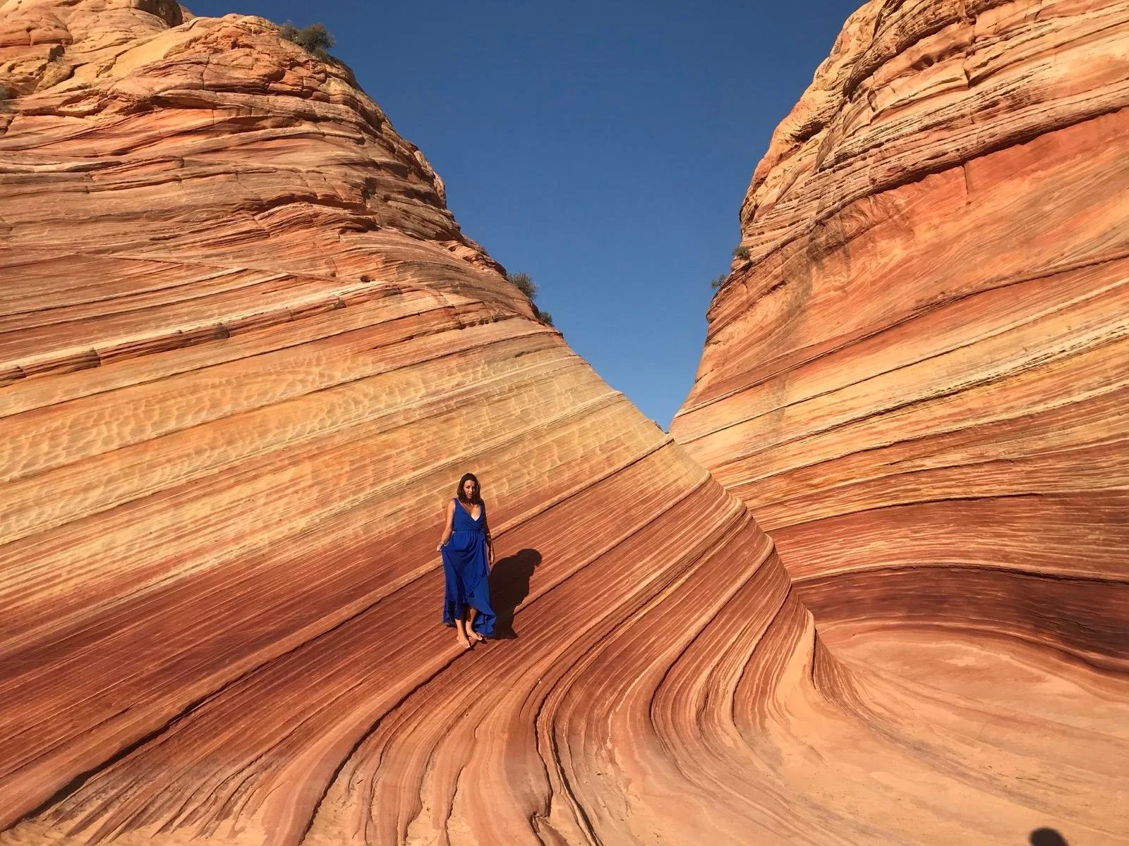 Une femme en robe bleue se tient sur des formations rocheuses en forme de vagues aux couleurs ocres et rouges sous un ciel bleu clair.
