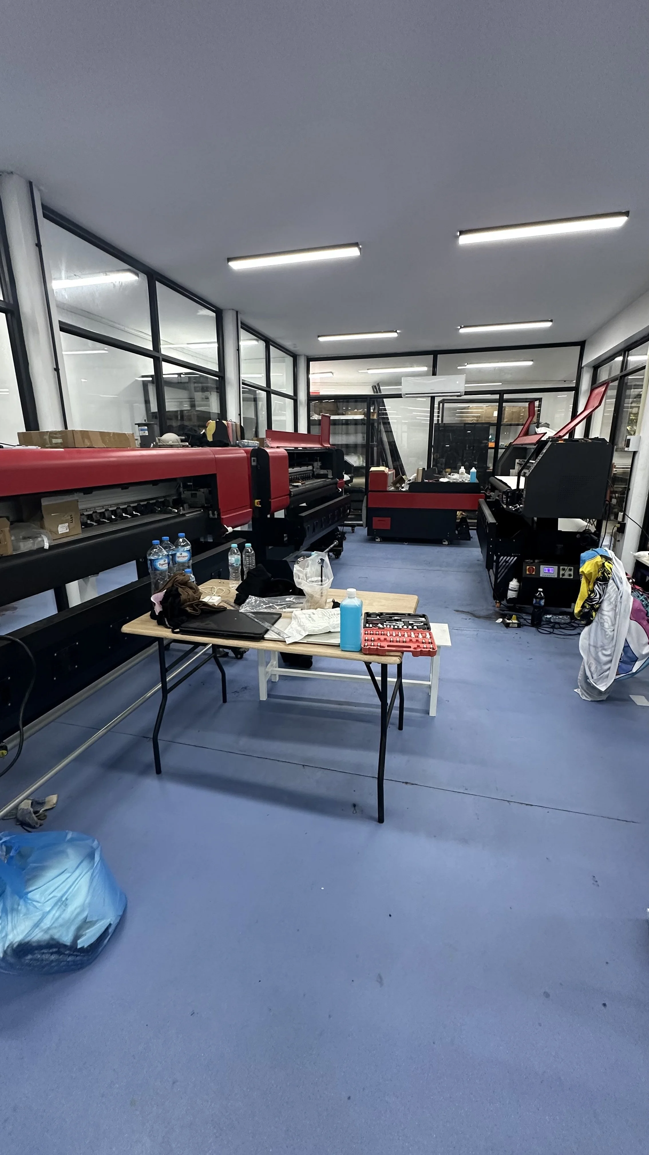 A room with large glass windows, containing industrial printing or manufacturing equipment, a table with tools, bottles, and miscellaneous items, and laundry or clothing on a chair.