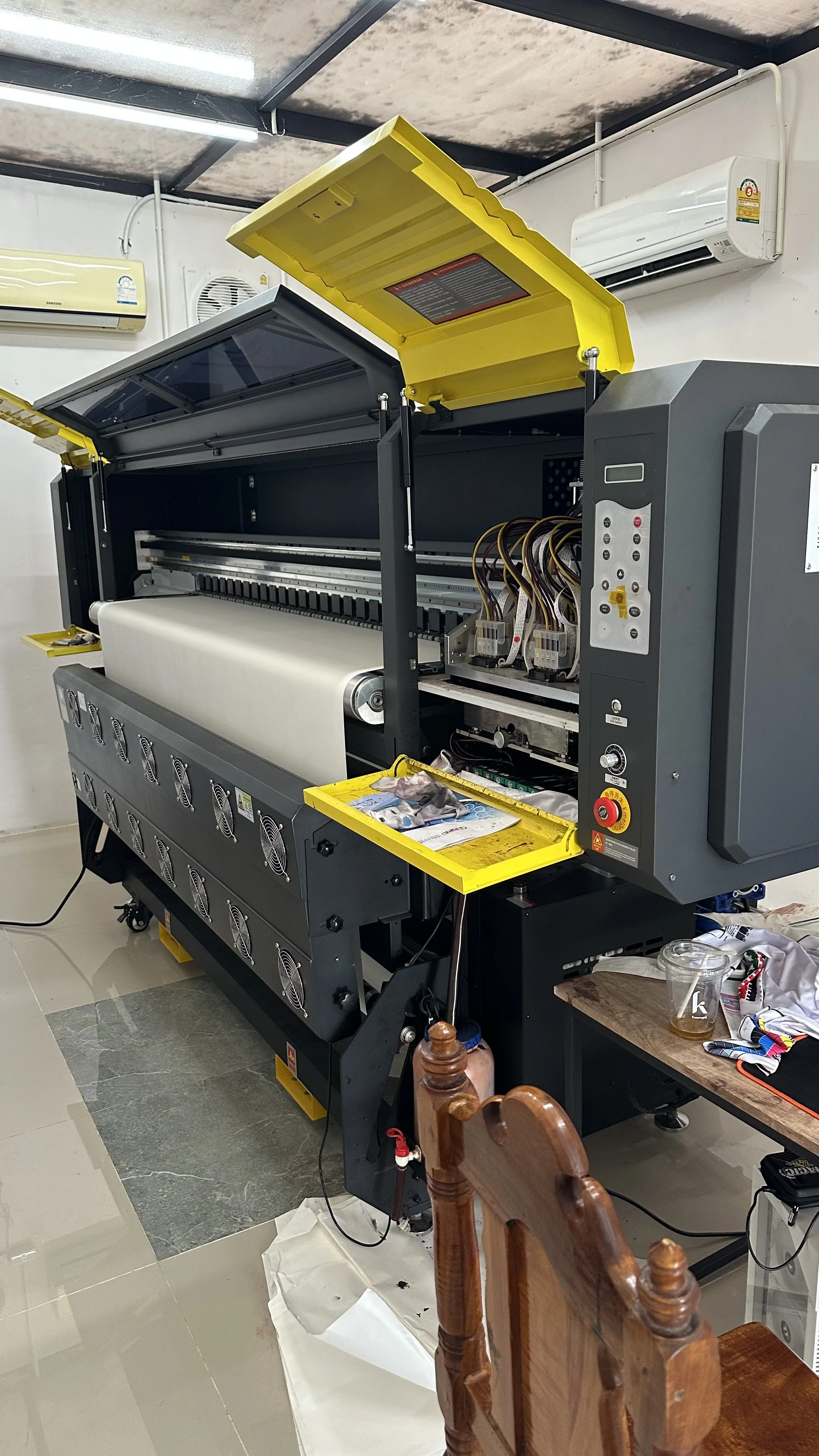 Open large industrial printer with yellow safety cover raised, showing interior components, in a room with air conditioning units and scattered items on a table and floor.