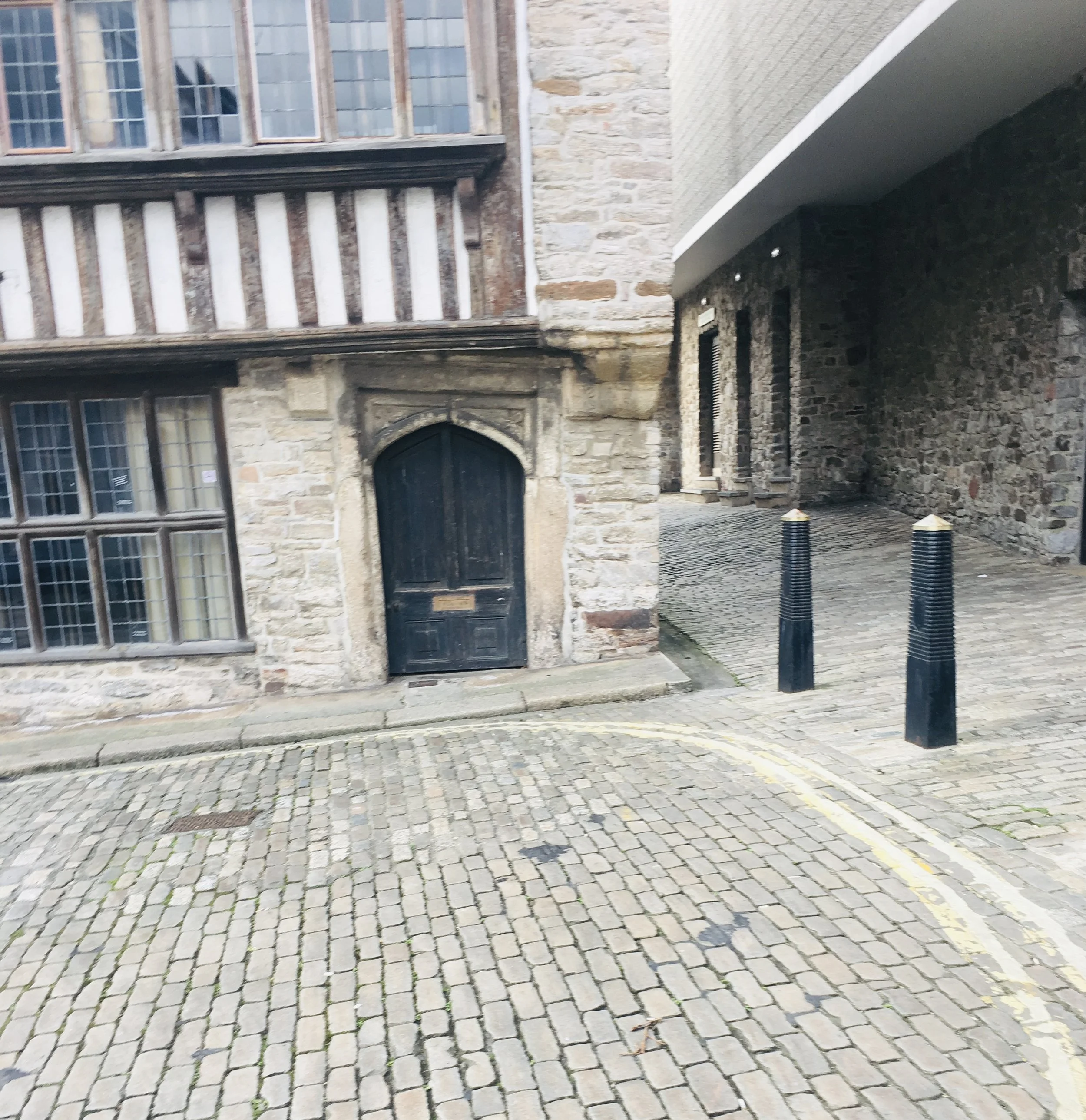 A stone and brick building with barred windows and a black door, cobblestone street, and black bollards.