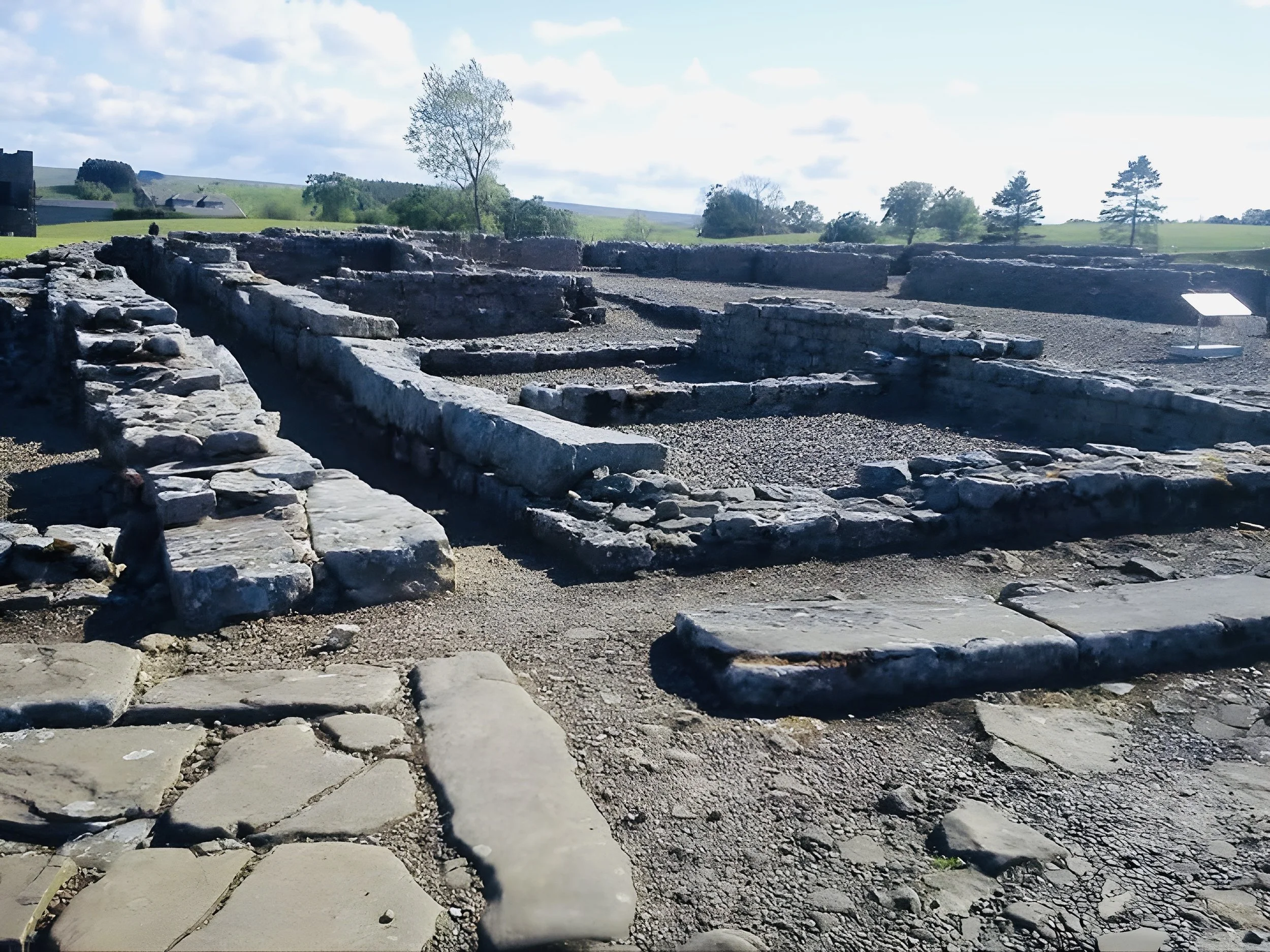 Ruins of an ancient stone structure with remnants of walls and foundations in an outdoor archaeological site, with green hills and trees in the background under a partly cloudy sky.