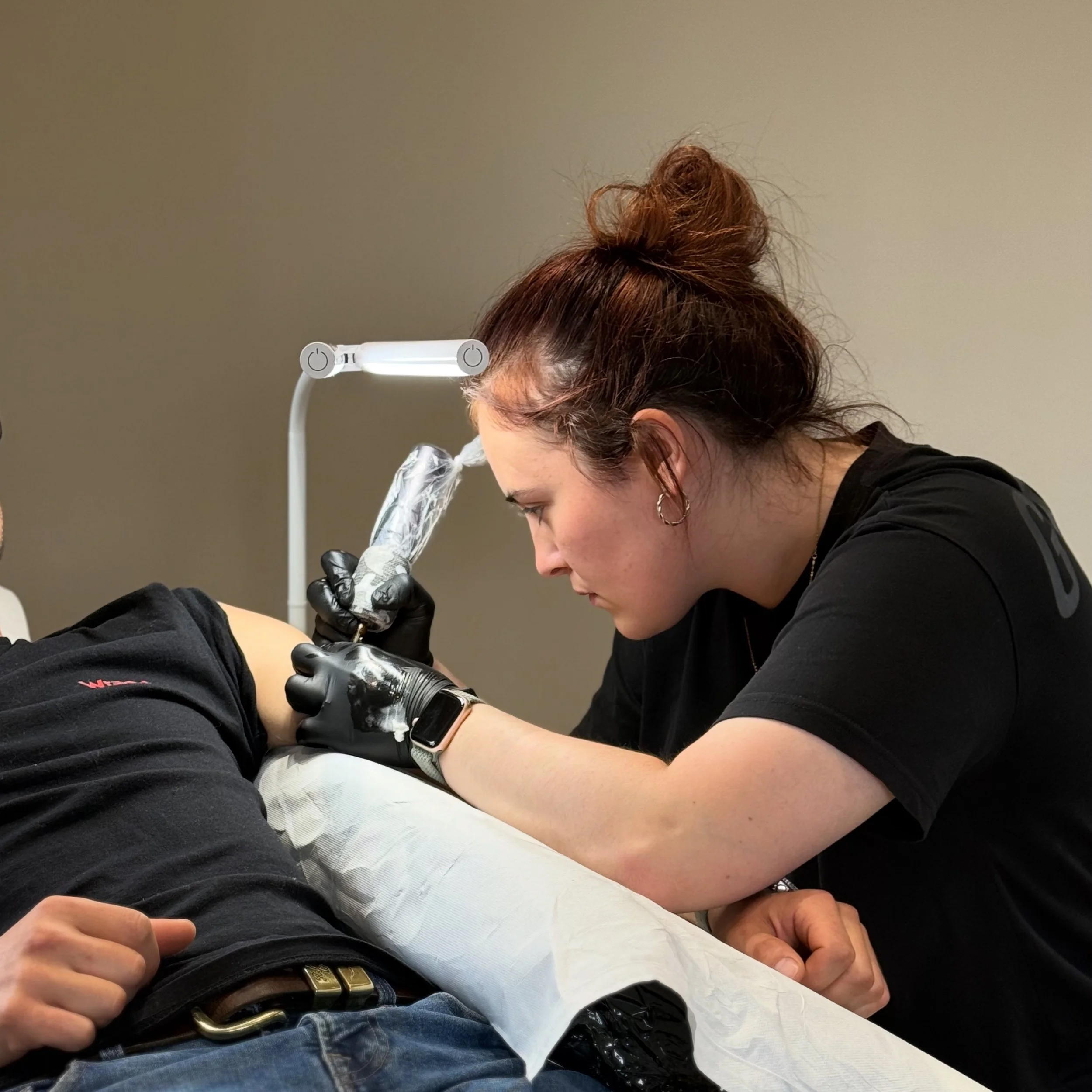 A person with red hair styled in a bun, wearing a black shirt is tattooing someone's arm. The process is taking place indoors with a beige wall in the background.