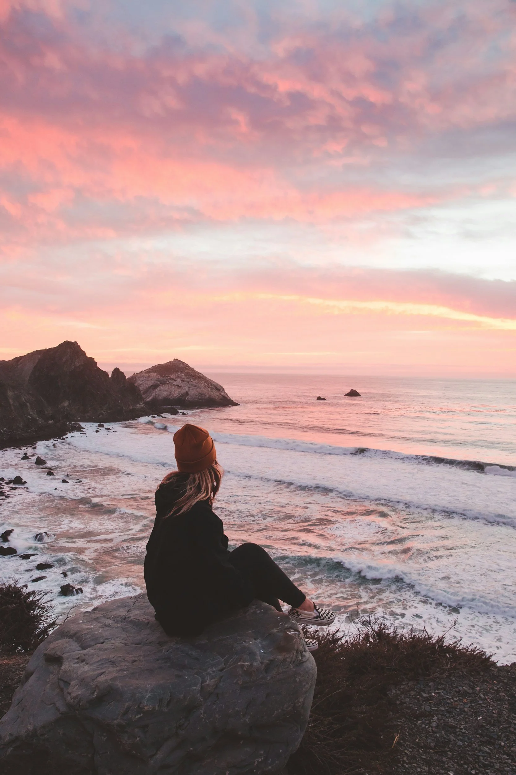 A person sitting on a large rock by the ocean during sunset, wearing a brown beanie, black jacket, and checkered shoes, looking at pink and purple clouds in the sky.