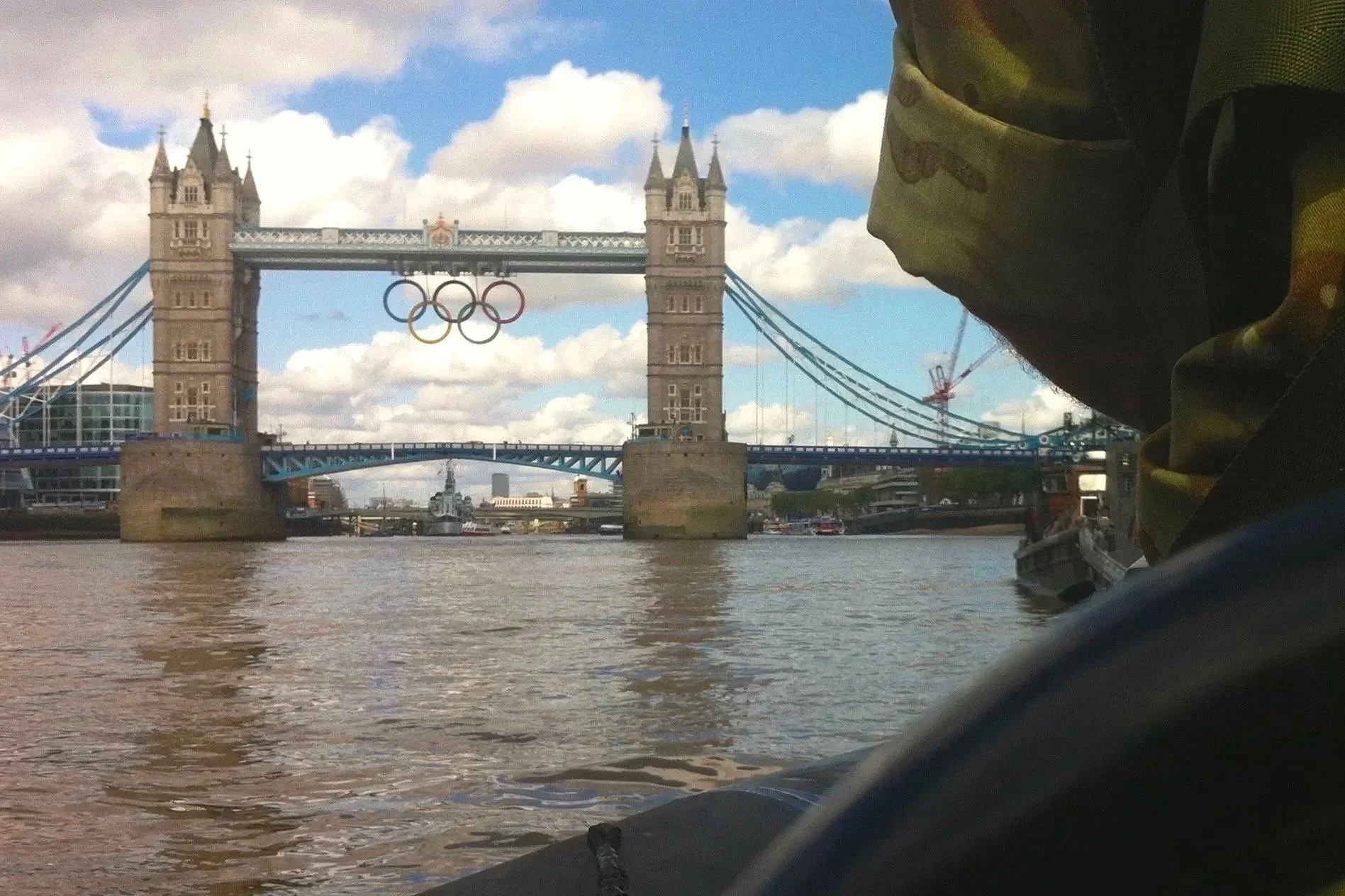 The London Tower Bridge spans the River Thames with the Olympic rings hanging from the top. The sky is partly cloudy, and buildings can be seen in the background.