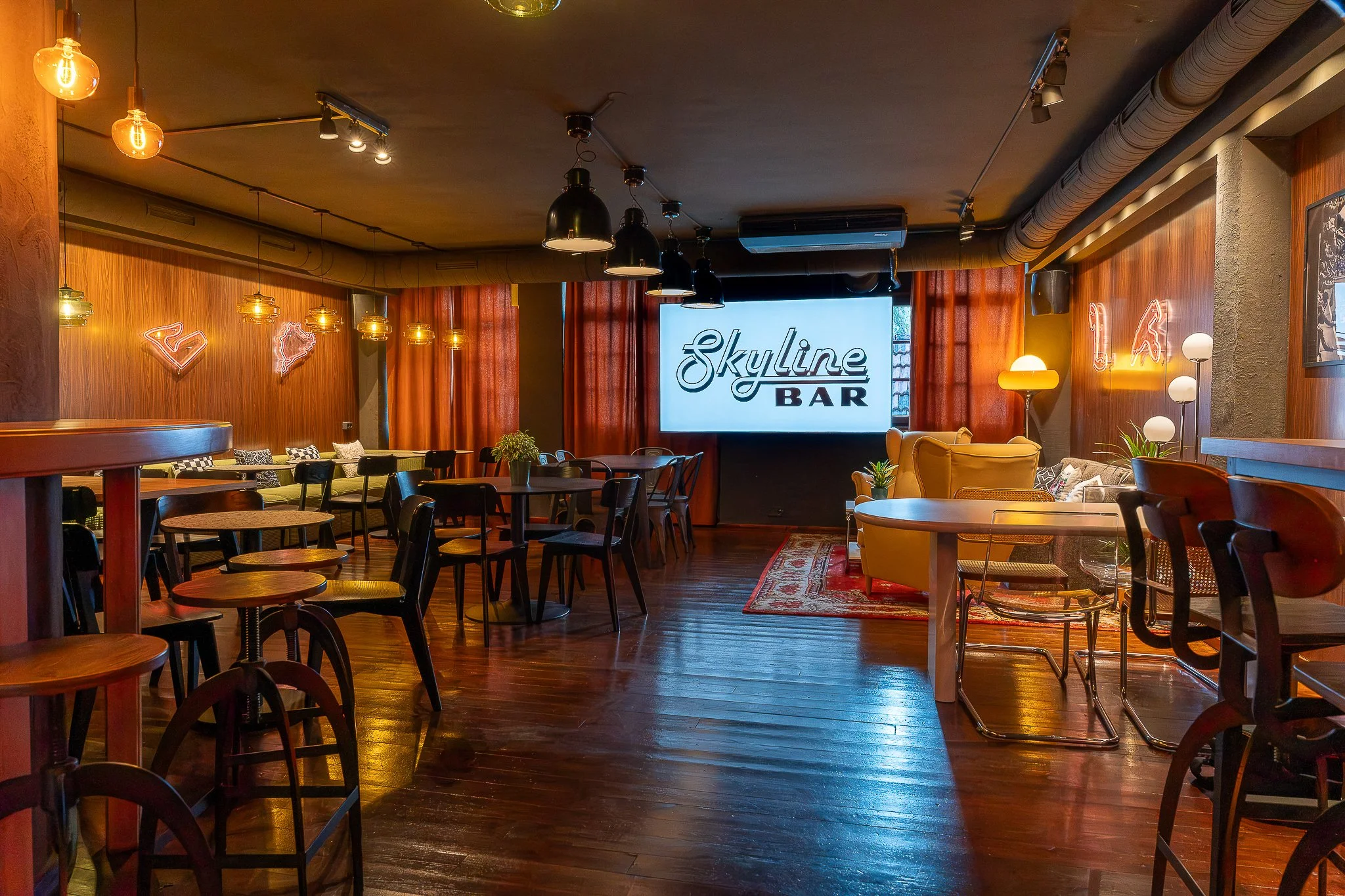 Interior of a bar or lounge with wooden floors and walls, warm lighting, various chairs and tables, and a large illuminated sign that reads 'Skyline BAR', decorated with neon-style light art and plants.
