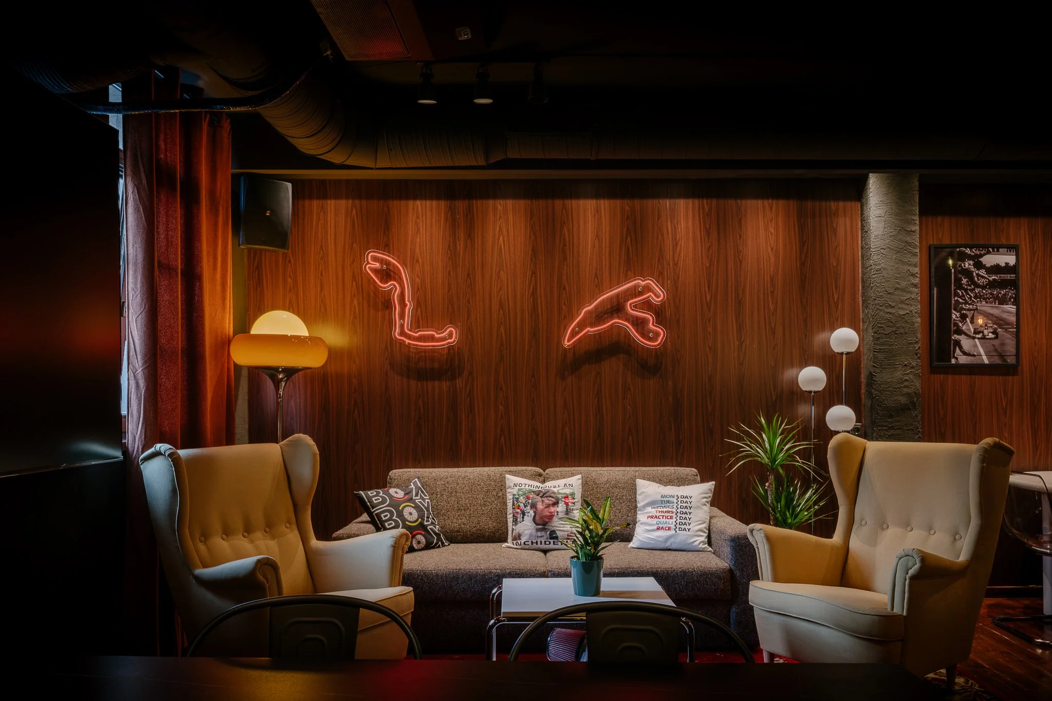 A cozy living room with beige armchairs, a sofa with throw pillows, a small coffee table with a plant, warm lighting including a yellow lamp, and decorative neon lights on a wood-paneled wall.
