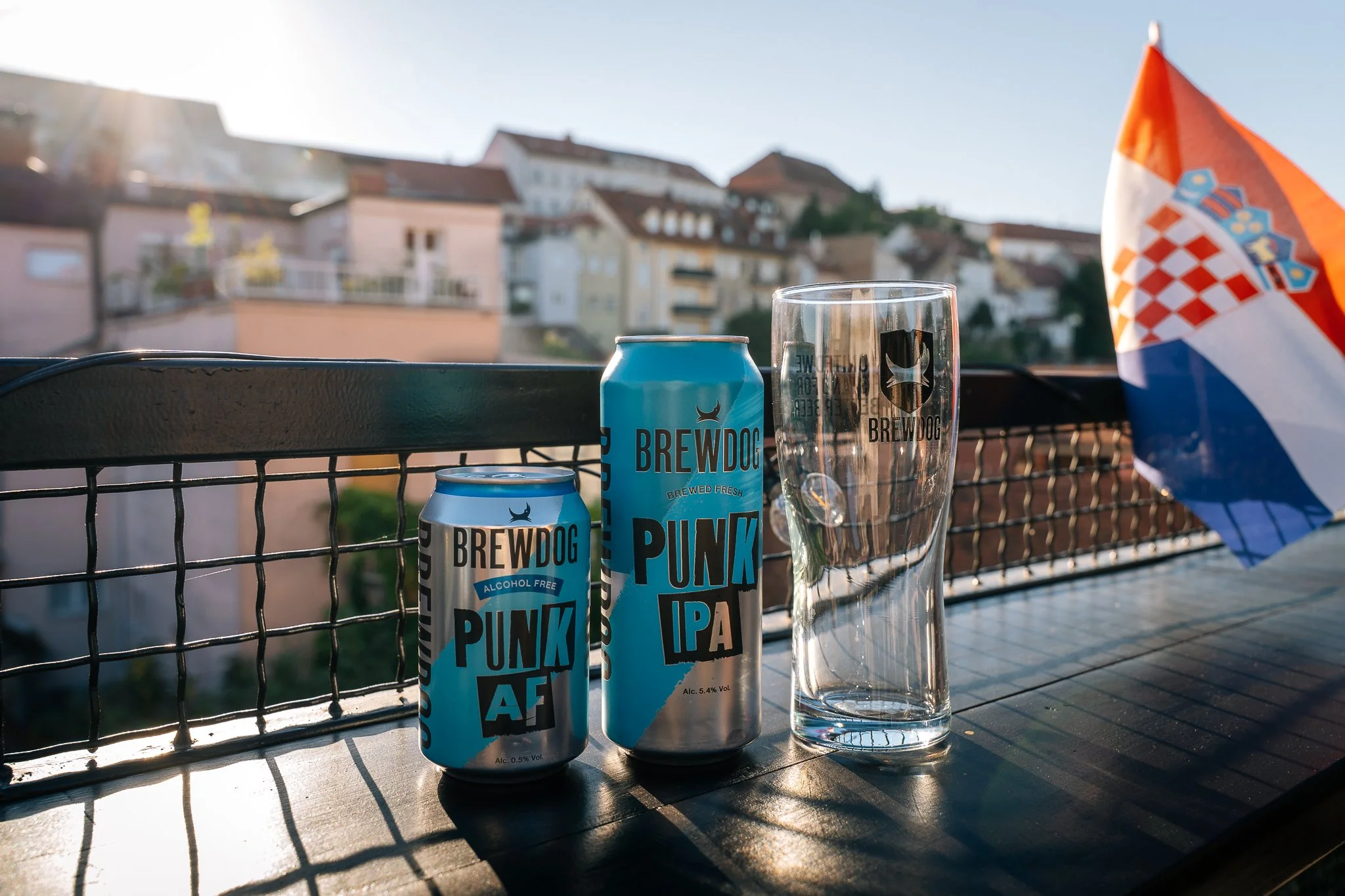 On a balcony, there are two cans and one glass of Brewdog non-alcoholic craft beer, with a Croatian flag on the side. The background shows a hillside neighborhood with houses and trees, in sunset light.