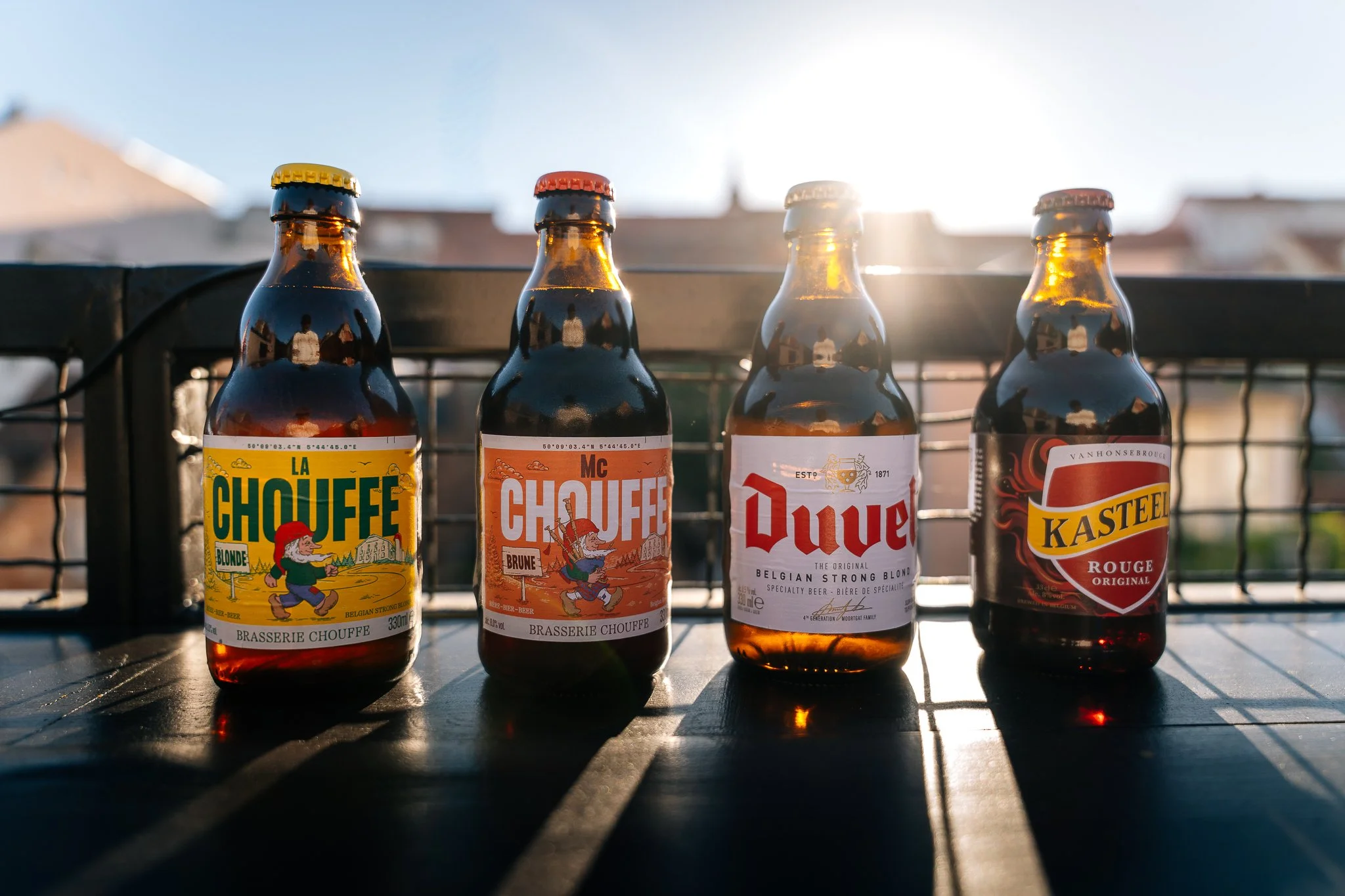 Four brown glass beer bottles with colorful labels standing on a black surface outdoors, with the sun shining brightly in the background.