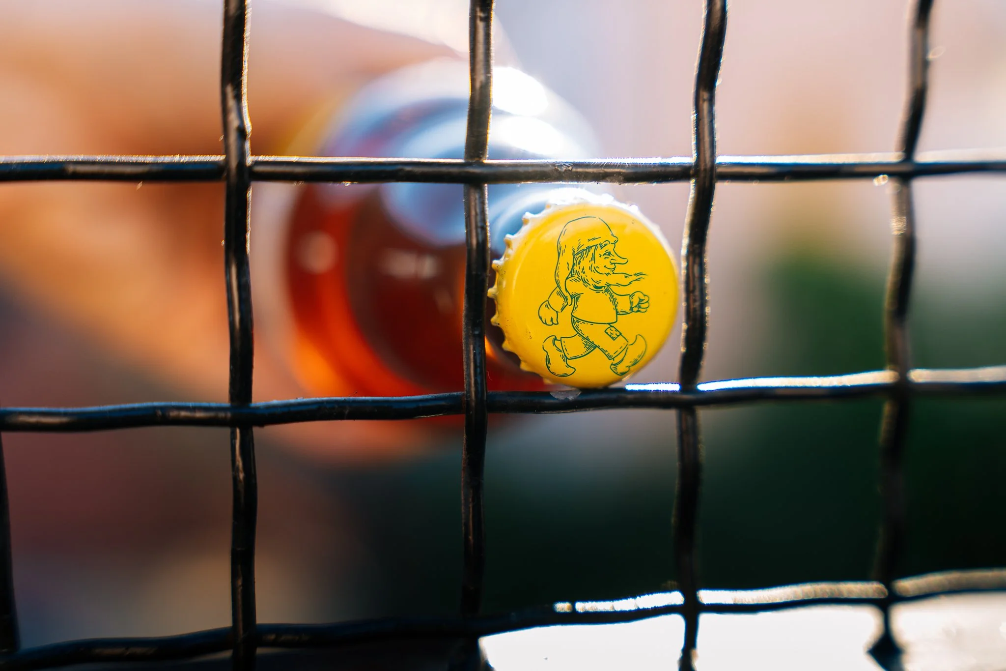 Close-up of a yellow bottle cap with a line drawing of a running girl, attached to a bottle, seen through a wire fence, with a blurry background.