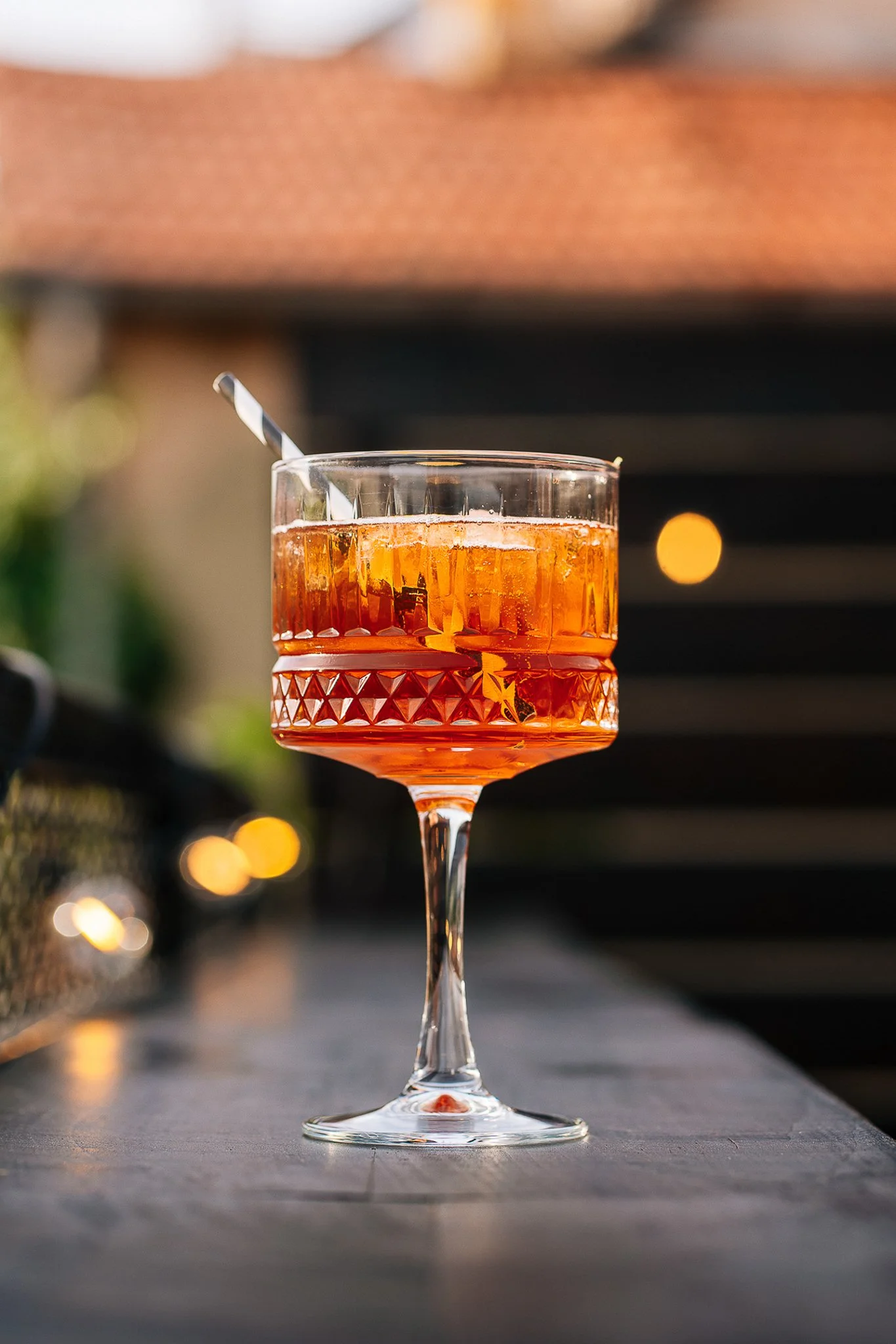 A glass of amber-colored cocktail with ice, served in a decorative stemmed glass with a striped straw, on a dark outdoor surface with blurred hanging lights in the background.
