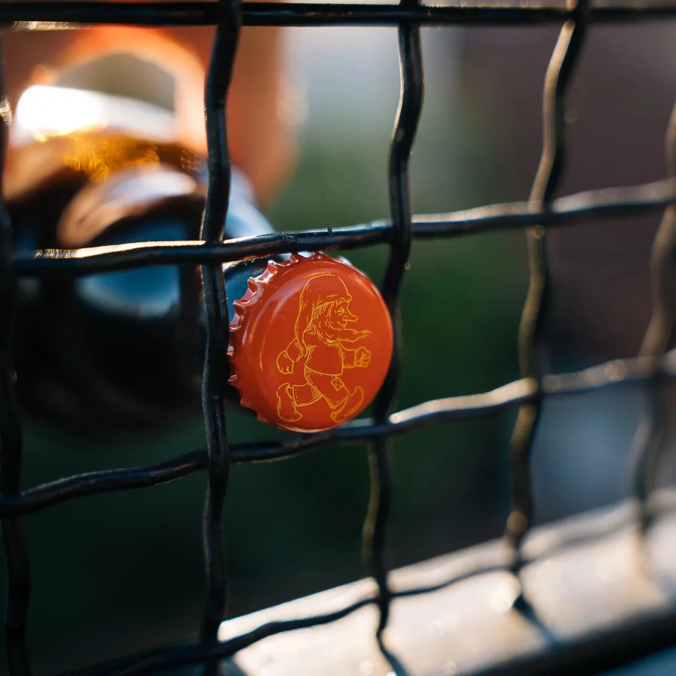 Close-up of a bottle cap with a cartoon gnome, seen through a wire fence, with sunlight illuminating the scene.
