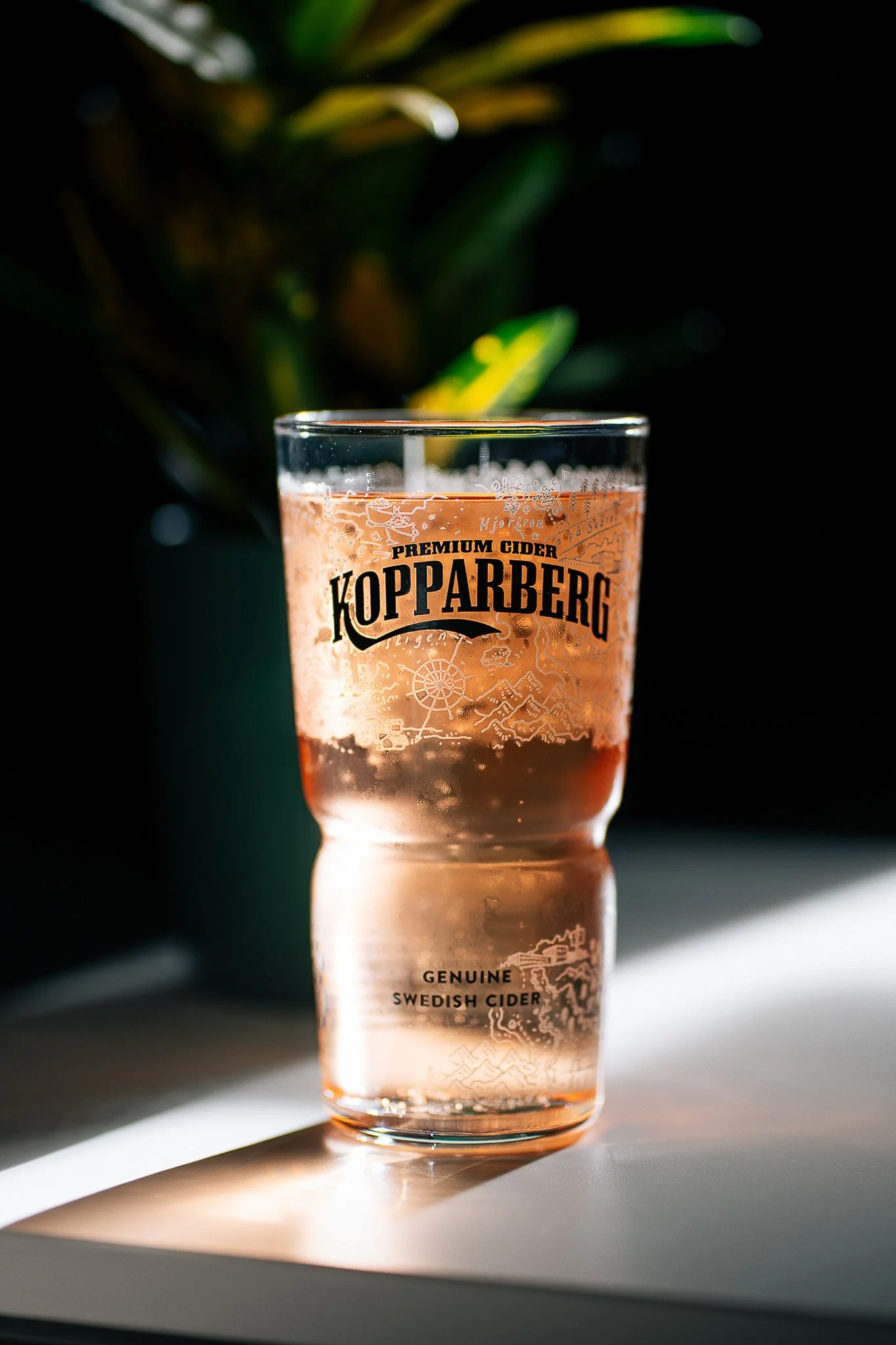 A glass of pink iced cider with a label that reads 'Kopparberg Premium Cider, Genuine Swedish Cider' placed on a white surface with shadows and sunlight, with a blurred background that includes green leaves.