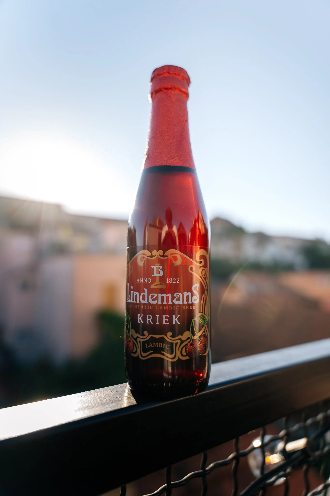 A bottle of Lindemans Kriek Lambic beer on a balcony rail, with blurred buildings and a blue sky in the background.