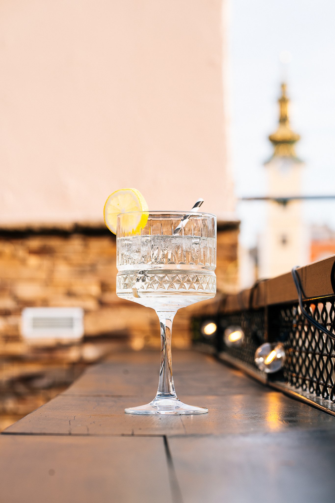 A glass of water with a lemon slice and a striped straw on a bar counter, with a blurry outdoor scene in the background.