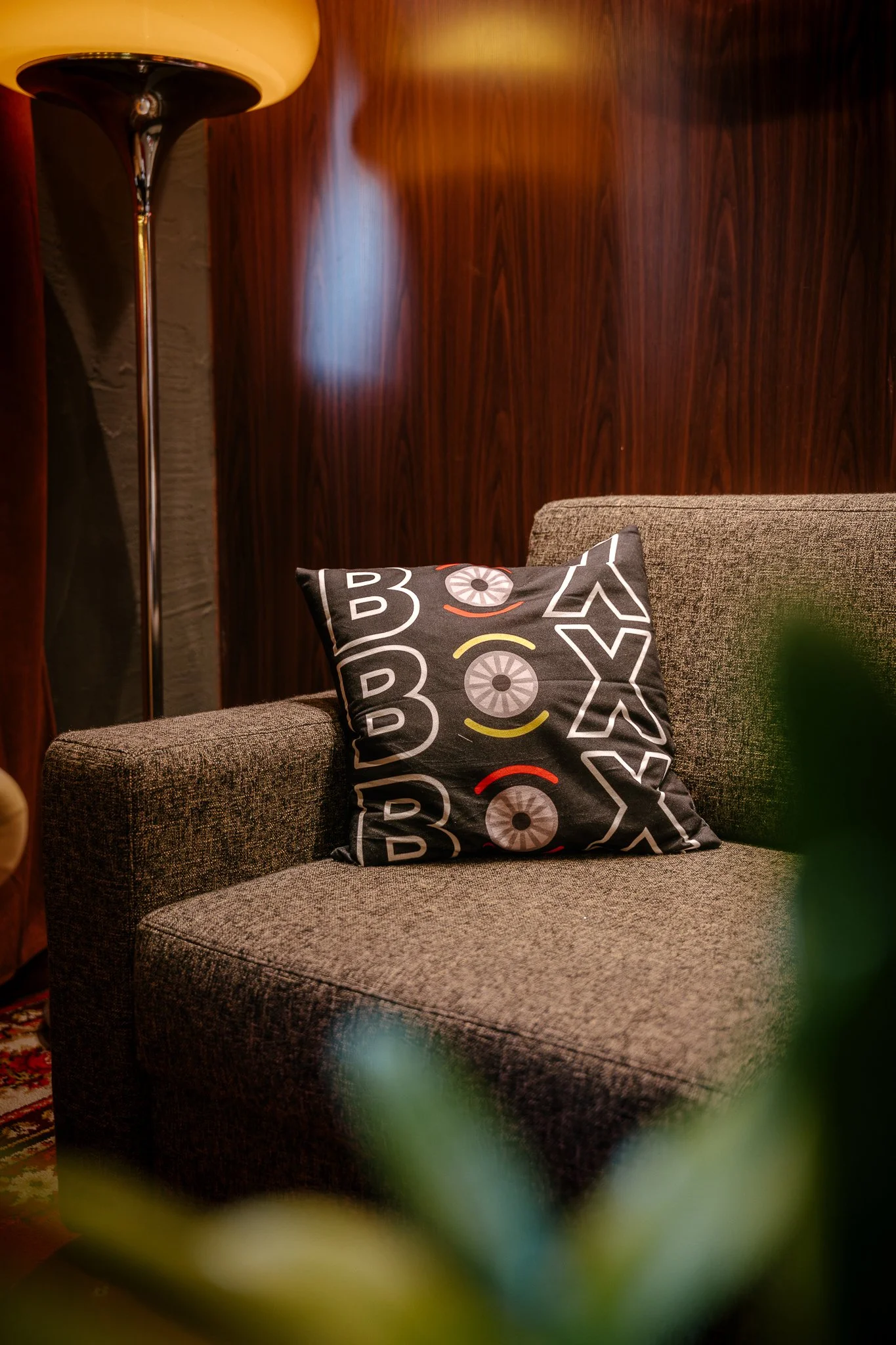 A cozy indoor scene featuring a brown textured sofa with a decorative black pillow that has colorful abstract patterns. Behind the sofa is a dark wooden wall and a tall modern lamp with a beige shade. In the foreground, blurred green leaves frame the scene.