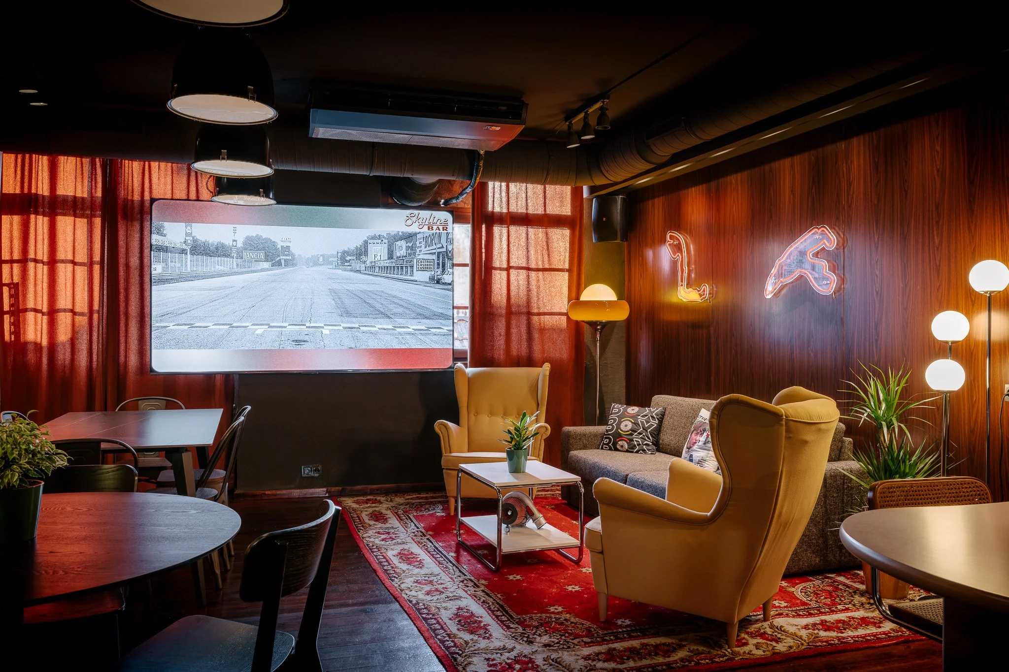 Interior of a cozy cafe with vintage furniture, a patterned rug, and a wooden accent wall adorned with neon signs of a whale and a dolphin, illuminated by modern lamps. Large windows with orange curtains reveal a track and field scene outside.