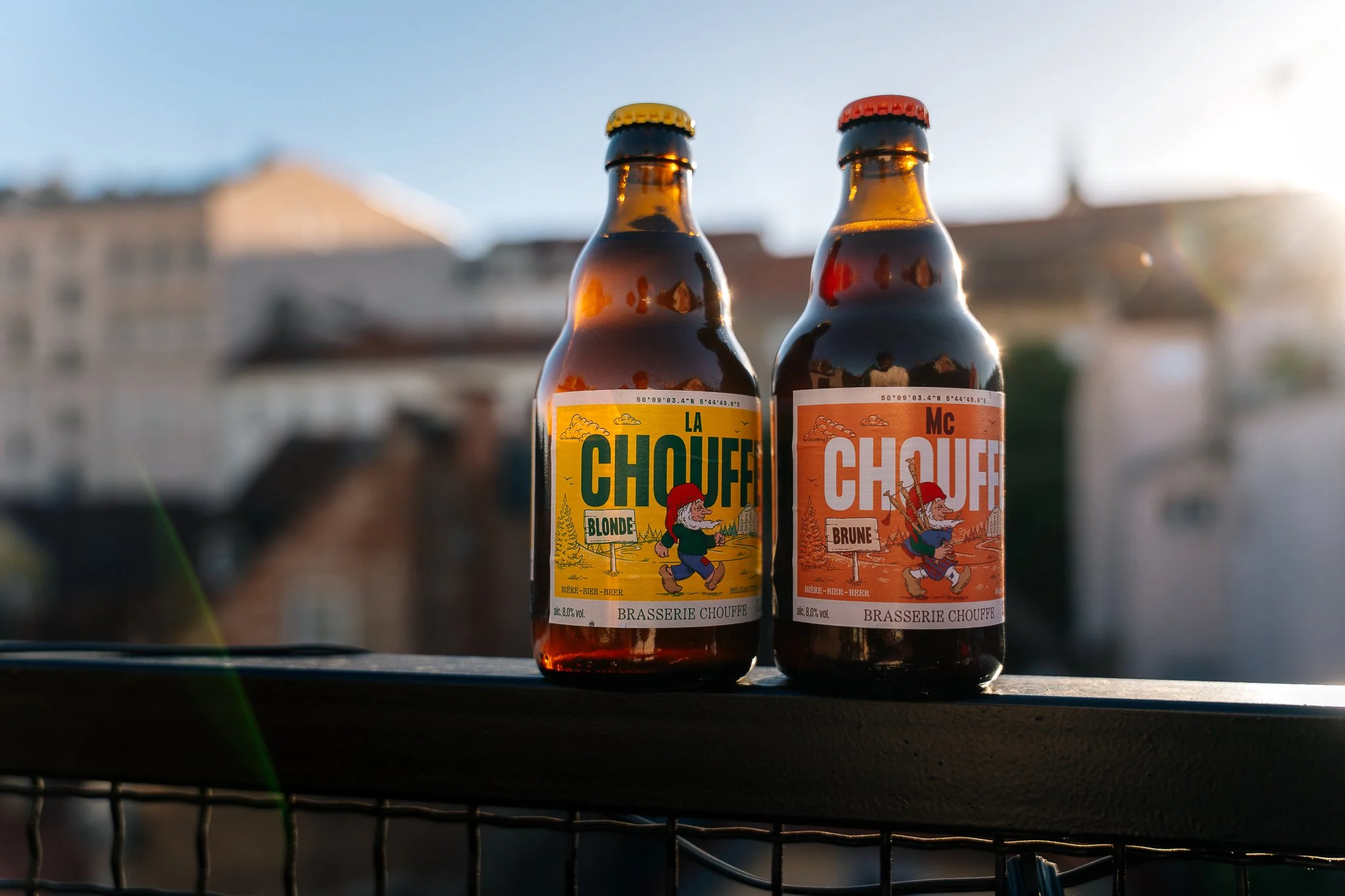 Two small bottles of beer with colorful labels, one yellow labeled 'La Chouffe Blonde' and one orange labeled 'MC Chouffe Brune,' sitting outdoors with a blurred cityscape in the background and sunlight shining on the bottles.