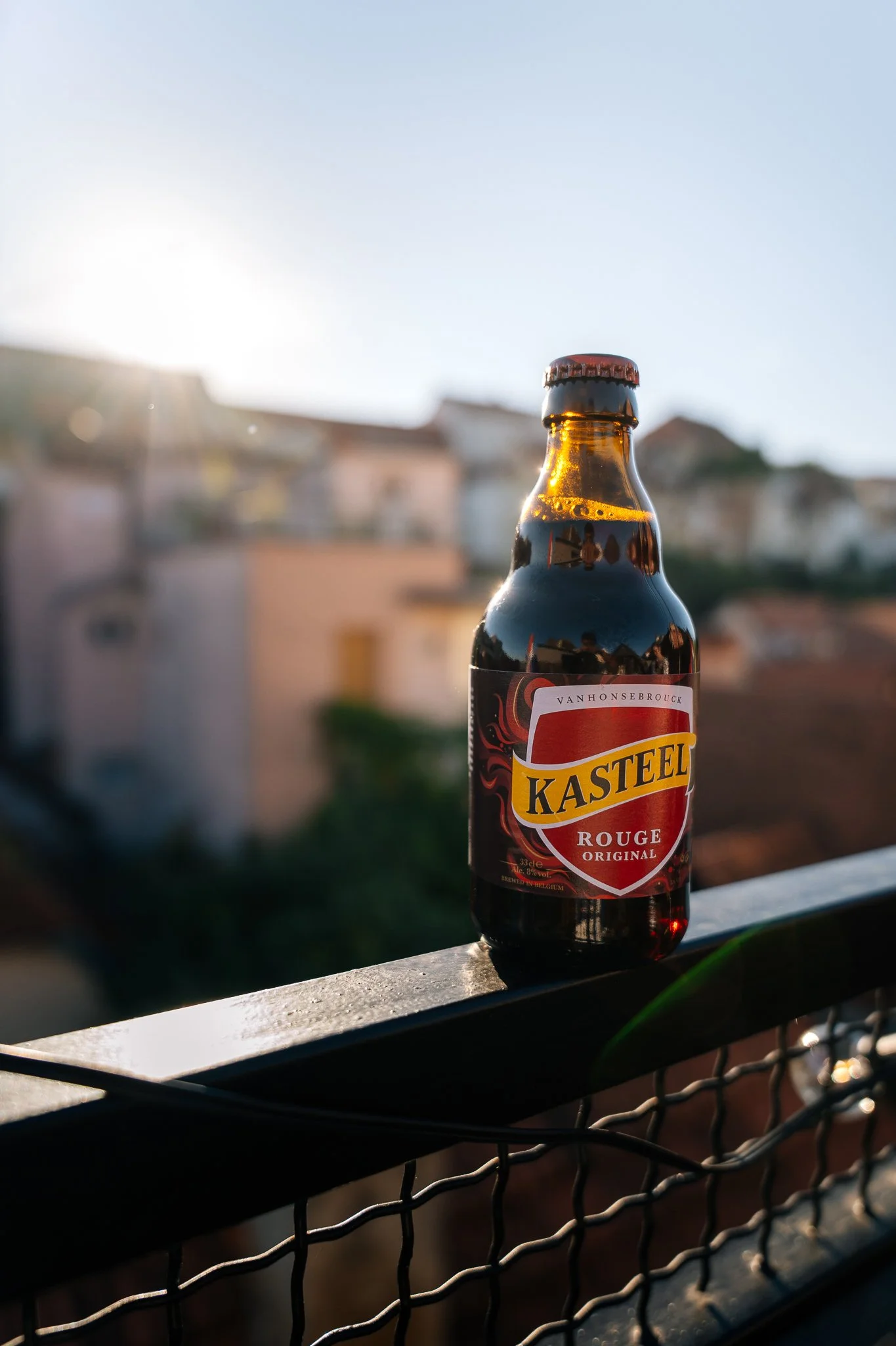 A bottle of Kasteel Rouge beer placed on a balcony railing with blurred houses in the background during sunset.