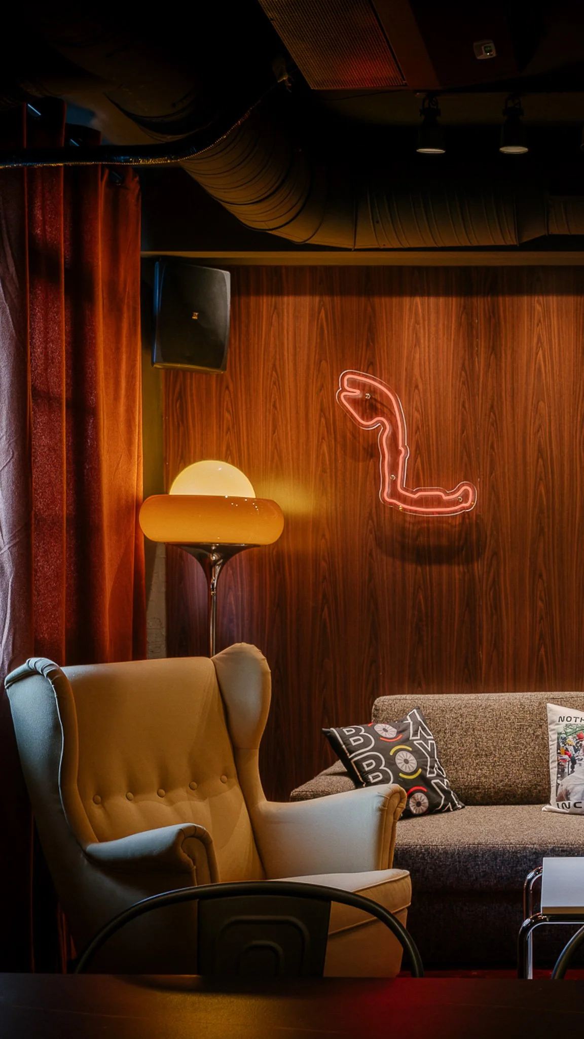 Interior of a cozy living room with a beige armchair, a sofa with patterned pillows, a tall lamp with a rounded shade, a wooden wall with a neon flamingo sign, a black speaker, and dark ceiling ductwork.