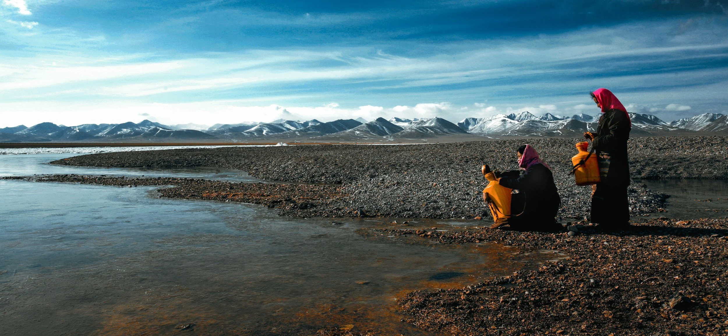 Zwei Frauen in Winterkleidung mit gelben Jerrys fischen im Fluss in einer bergigen, schneebedeckten Landschaft.