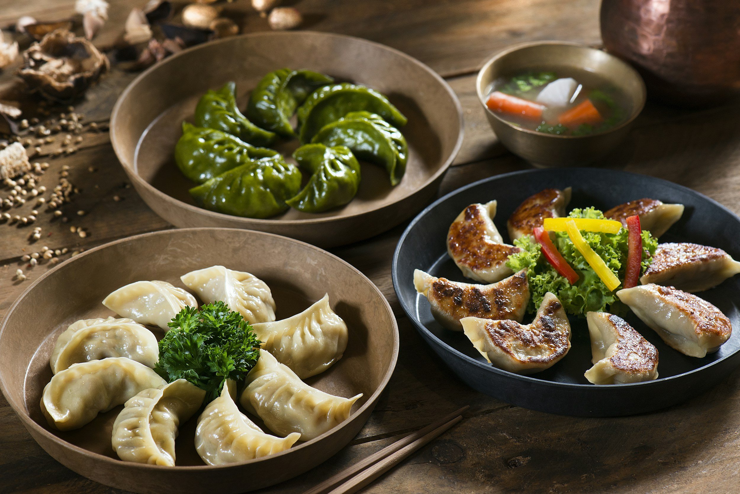 Verschiedene asiatische Gerichte, darunter gedämpfte Teigtaschen (Dimsum oder Gyoza), grüne Knödel, eine Suppe mit Karotten und Frühlingszwiebeln sowie eine Platte mit gebratenen Teigtaschen und Gemüse, auf einem dunklen Holztisch.