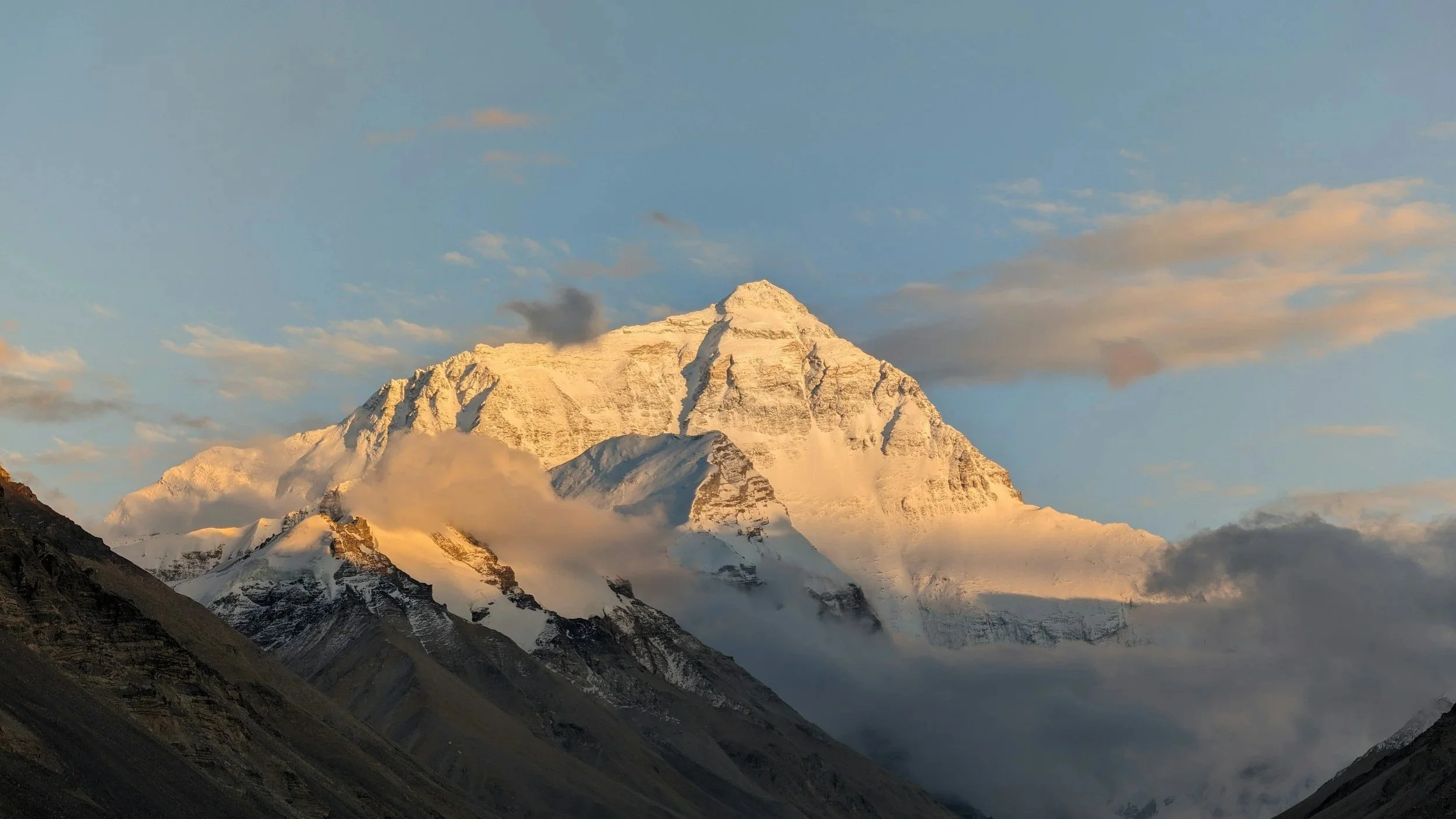 Berg mit schneebedecktem Gipfel in den Sonnenlichte, umgeben von Wolken und dunklen Hügeln im Vordergrund.