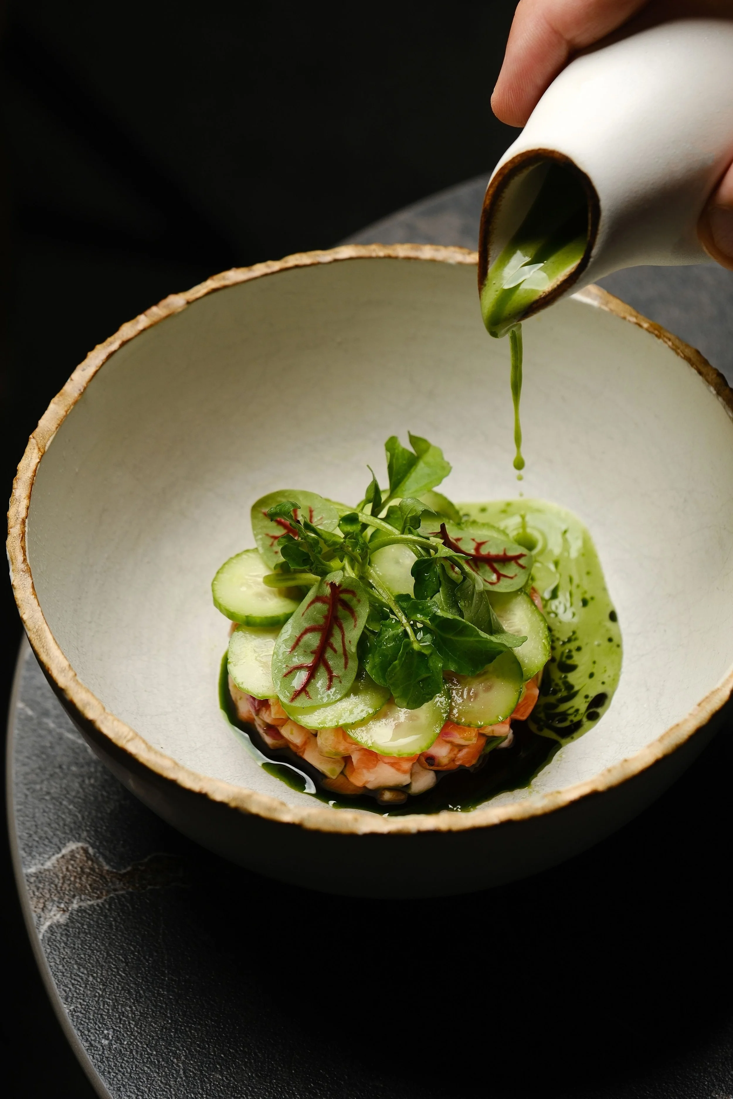 A dish of beautifully arranged food with sliced cucumbers, leafy greens, and a pinkish base, being dressed with a green sauce poured from a white container.