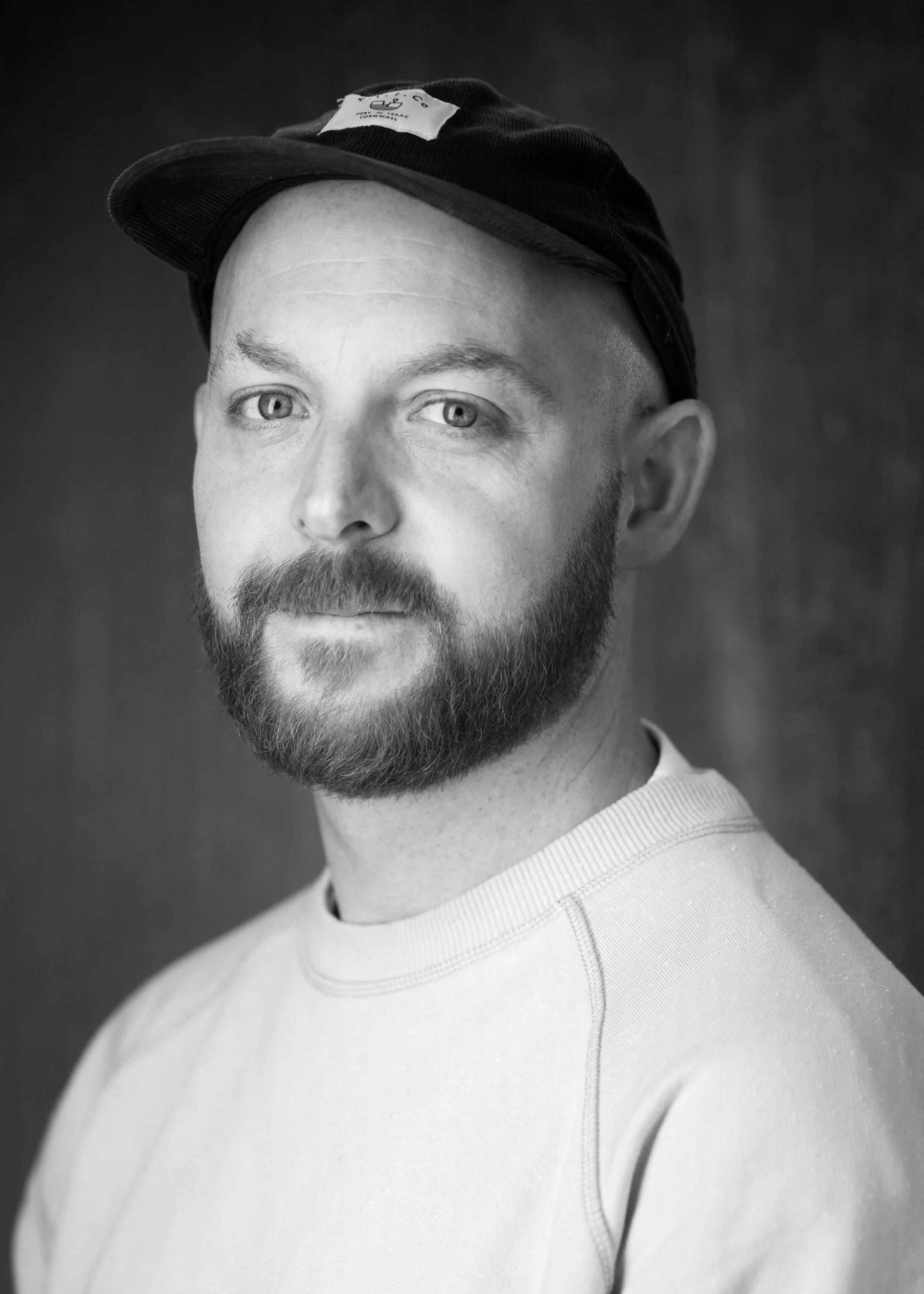 Black and white portrait of a man with a beard and mustache, wearing a baseball cap and a light-colored thermal shirt, looking directly at the camera.