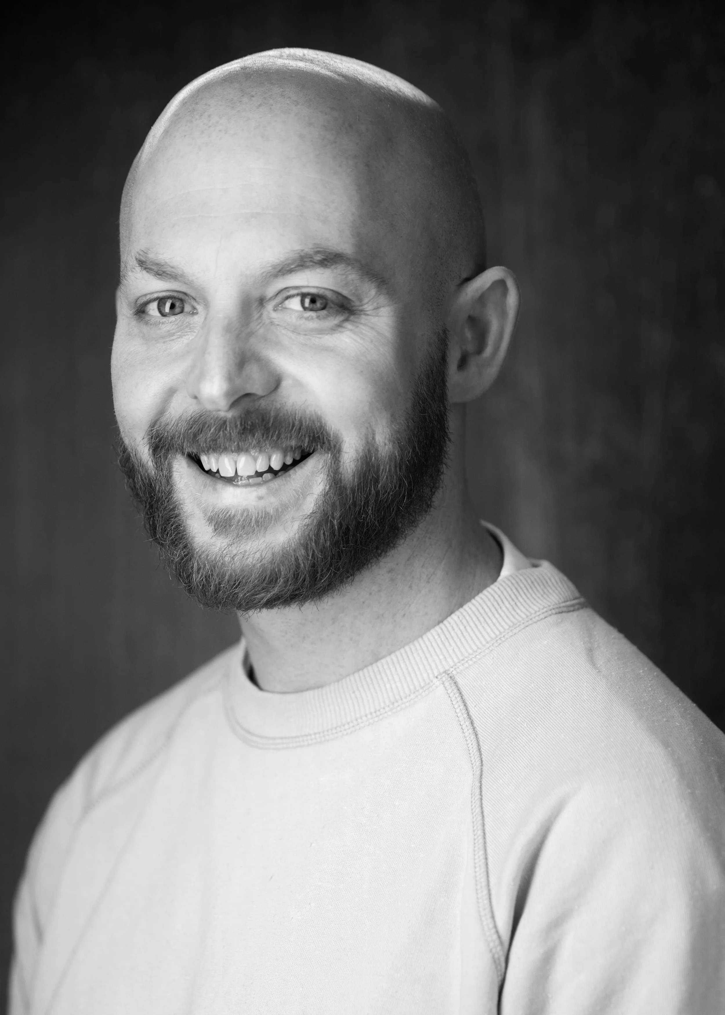 Black and white photo of a smiling man with a shaved head and beard, wearing a light-colored sweatshirt.