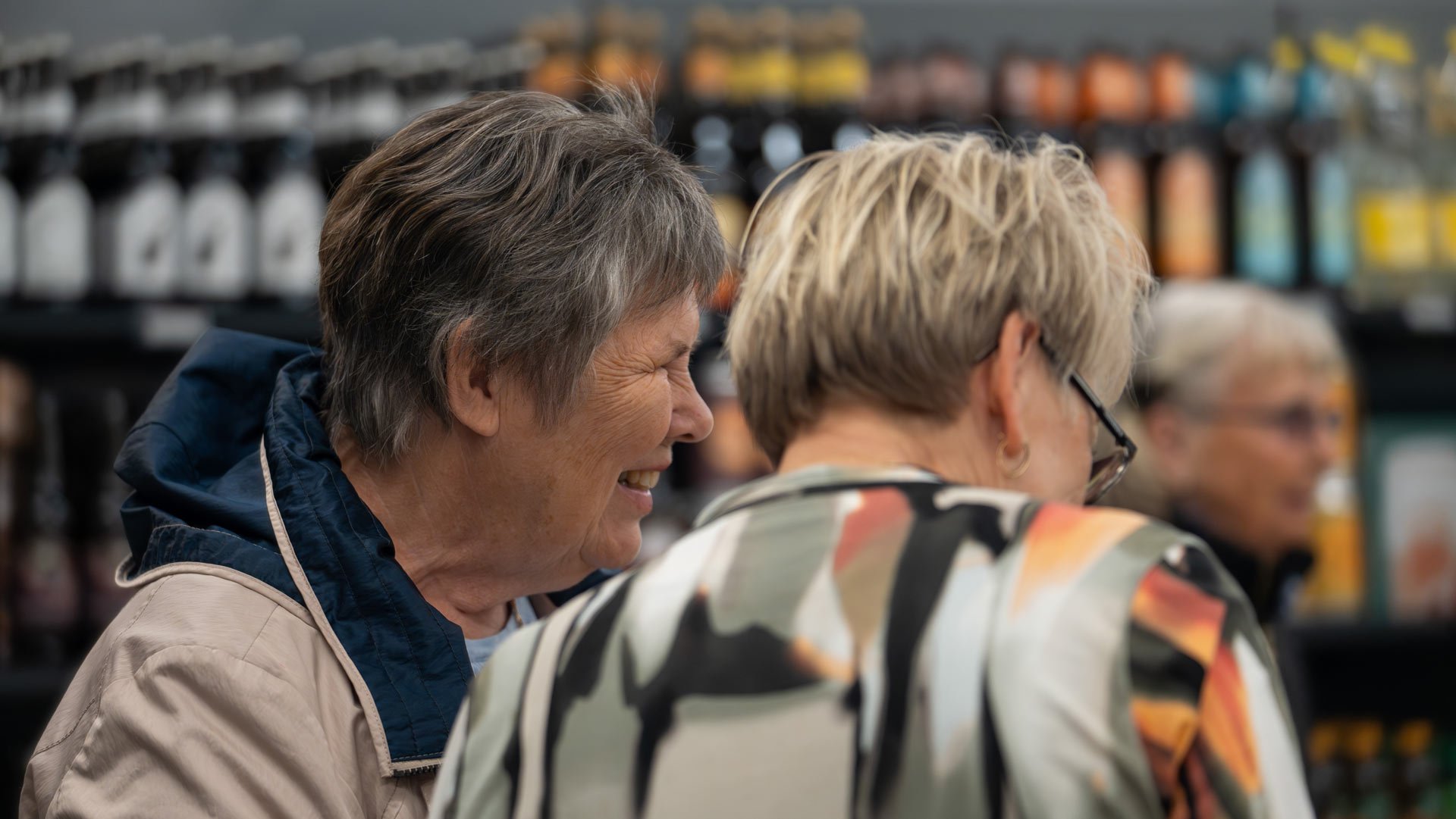 To kvinder står og snakker i en af vores butikker. Bag dem er der hylder med forskellige produkter.