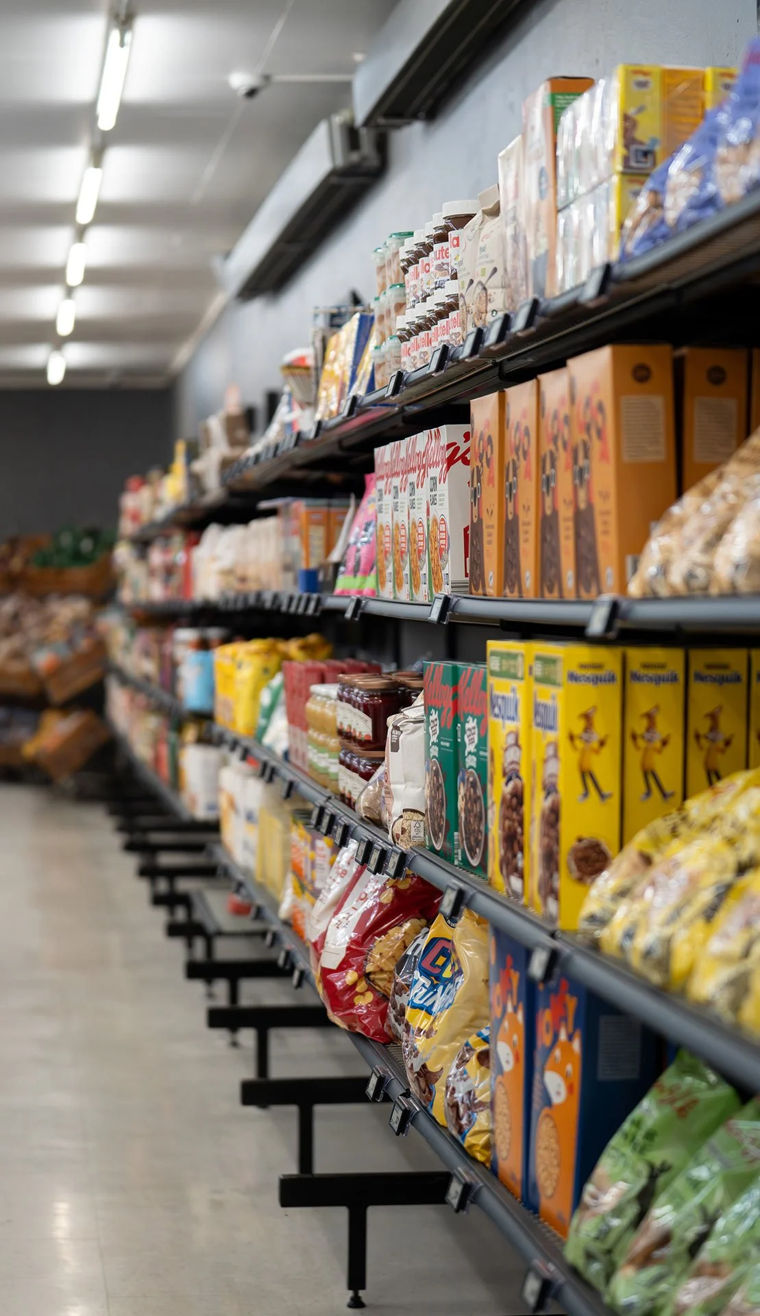 Billedet viser hylderne fra en af vores butikker fyldt med forskellige typer morgenmadsprodukter og snacks, herunder korn, knækbrød, chokolade, samt andre pakker.