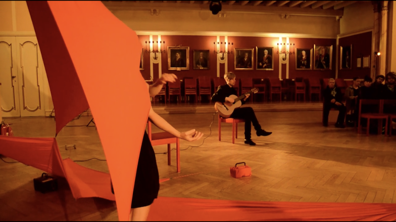 A person is performing with large red fabric in the foreground, while a person plays guitar in the background in a large, ornate room with paintings on the walls and an audience seated on red chairs.