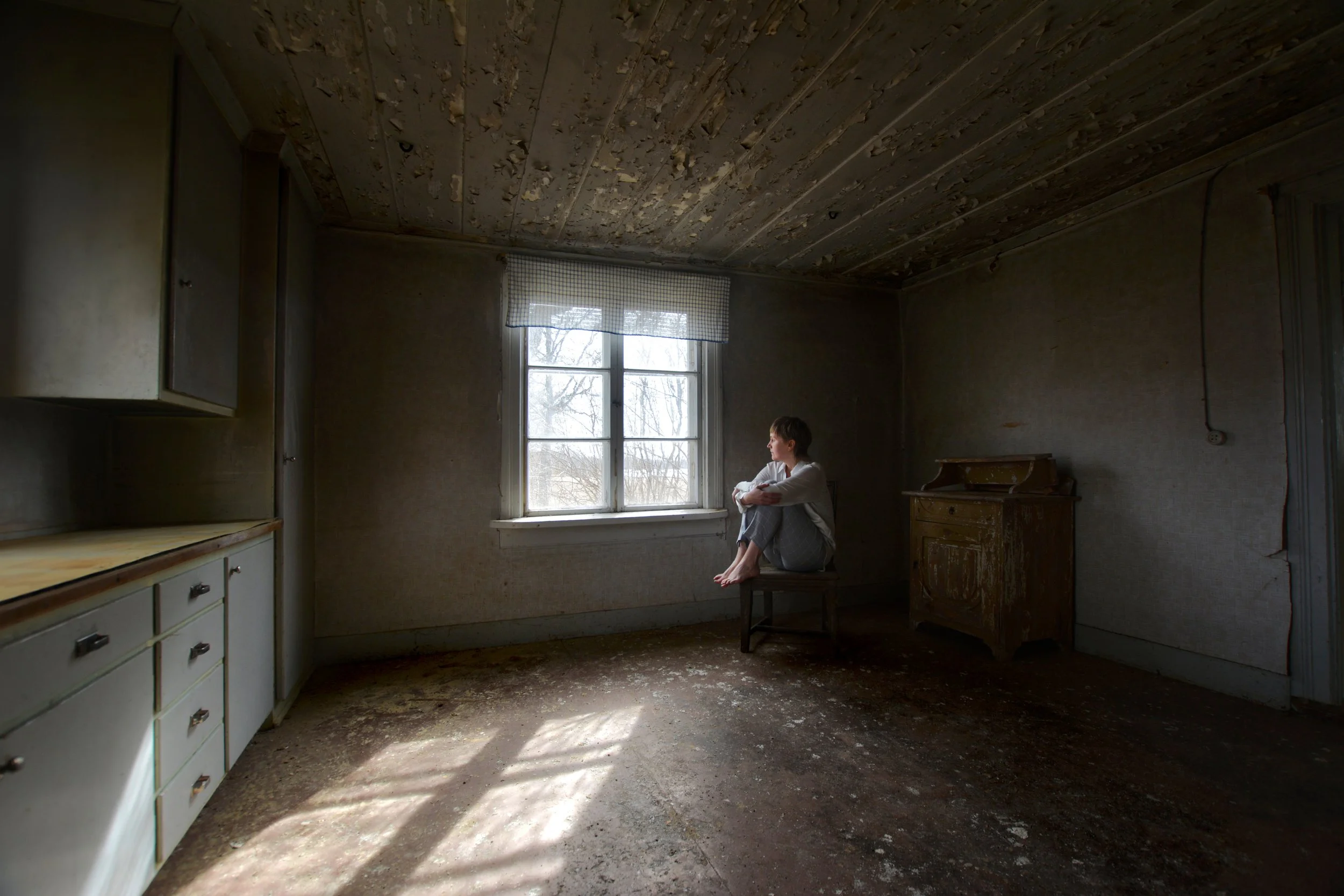 A woman sitting on a small table in an old, empty room with peeling wooden ceiling, dusty floor, and a window with natural light shining in.