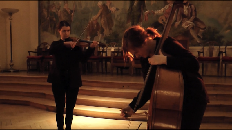 Two women playing musical instruments—one violin and one double bass—in a performance hall with a mural painting in the background.