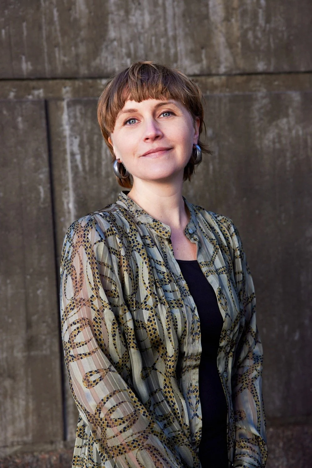 A woman with short brown hair and hoop earrings is standing in front of a concrete wall. She is wearing a patterned blouse over a black top and has a subtle smile on her face.