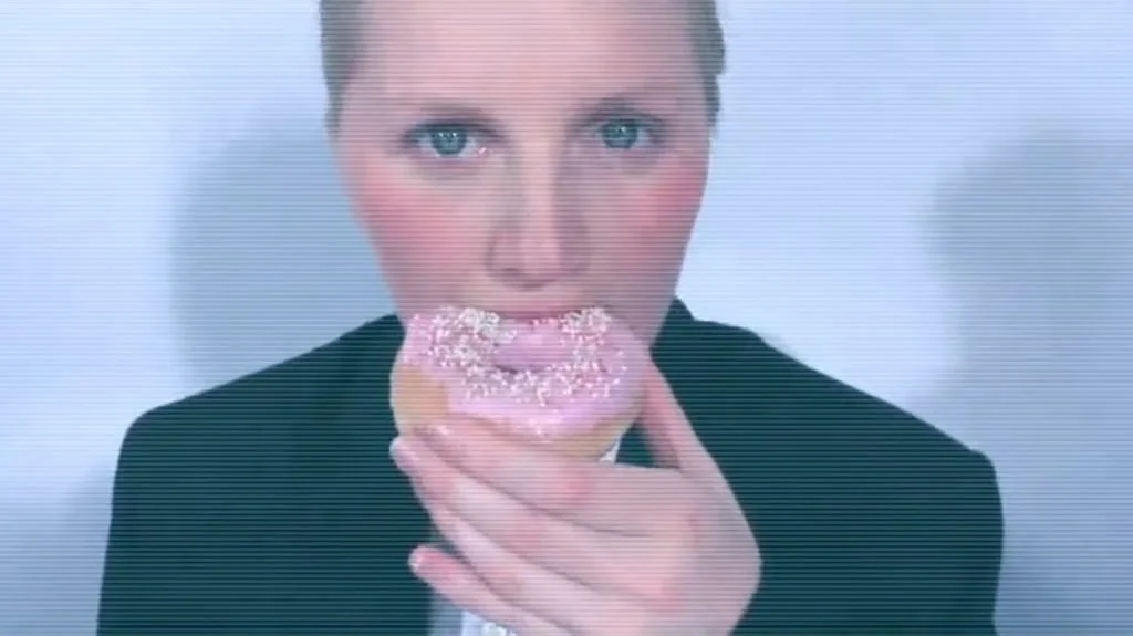 A woman with blonde hair and blue eyes holds a pink frosted donut with sprinkles close to her mouth, about to eat it.