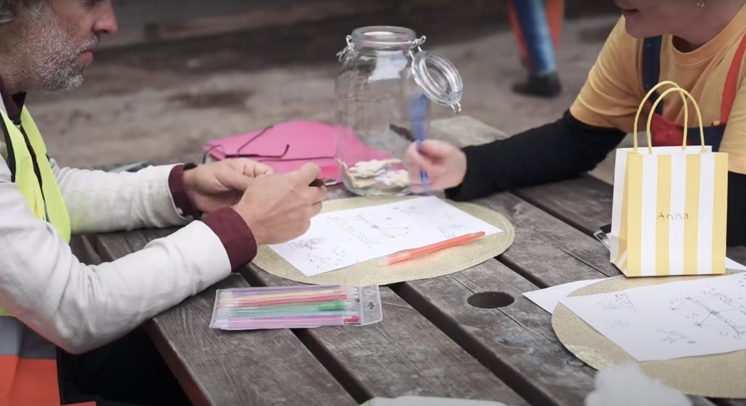 Two people at a outdoor table with papers, colorful pens, and a jar, engaged in a craft activity. One person is a man with gray hair and a beard wearing a white jacket with a maroon shirt underneath. The other person is a woman wearing an orange shir