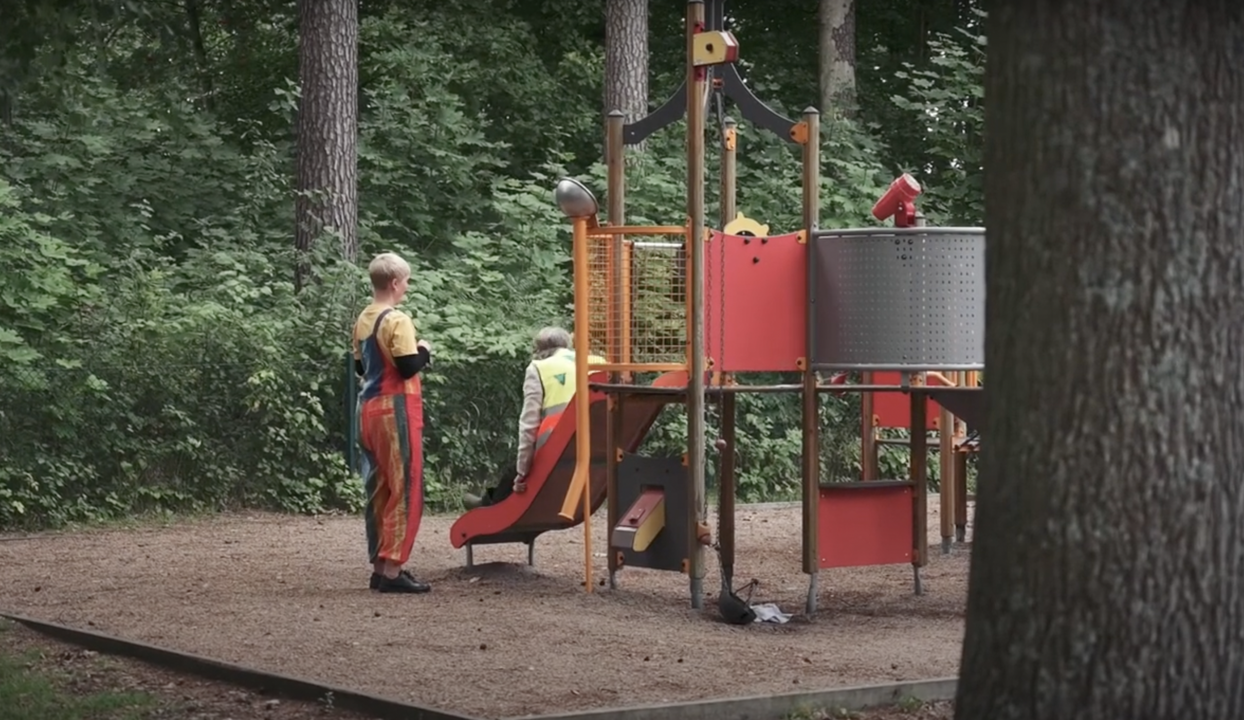 Two children at a playground surrounded by trees, with one child climbing a slide and the other looking on, wearing a safety vest.