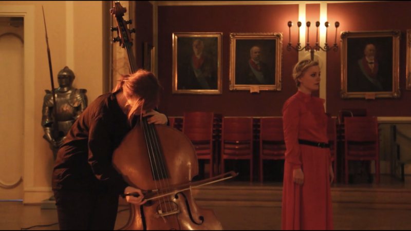 A woman standing on a stage in a red dress with a black belt, looking to the right. To her left, a person is playing a double bass, with their head resting on the instrument. The background features framed paintings, a chandelier, and a row of chairs