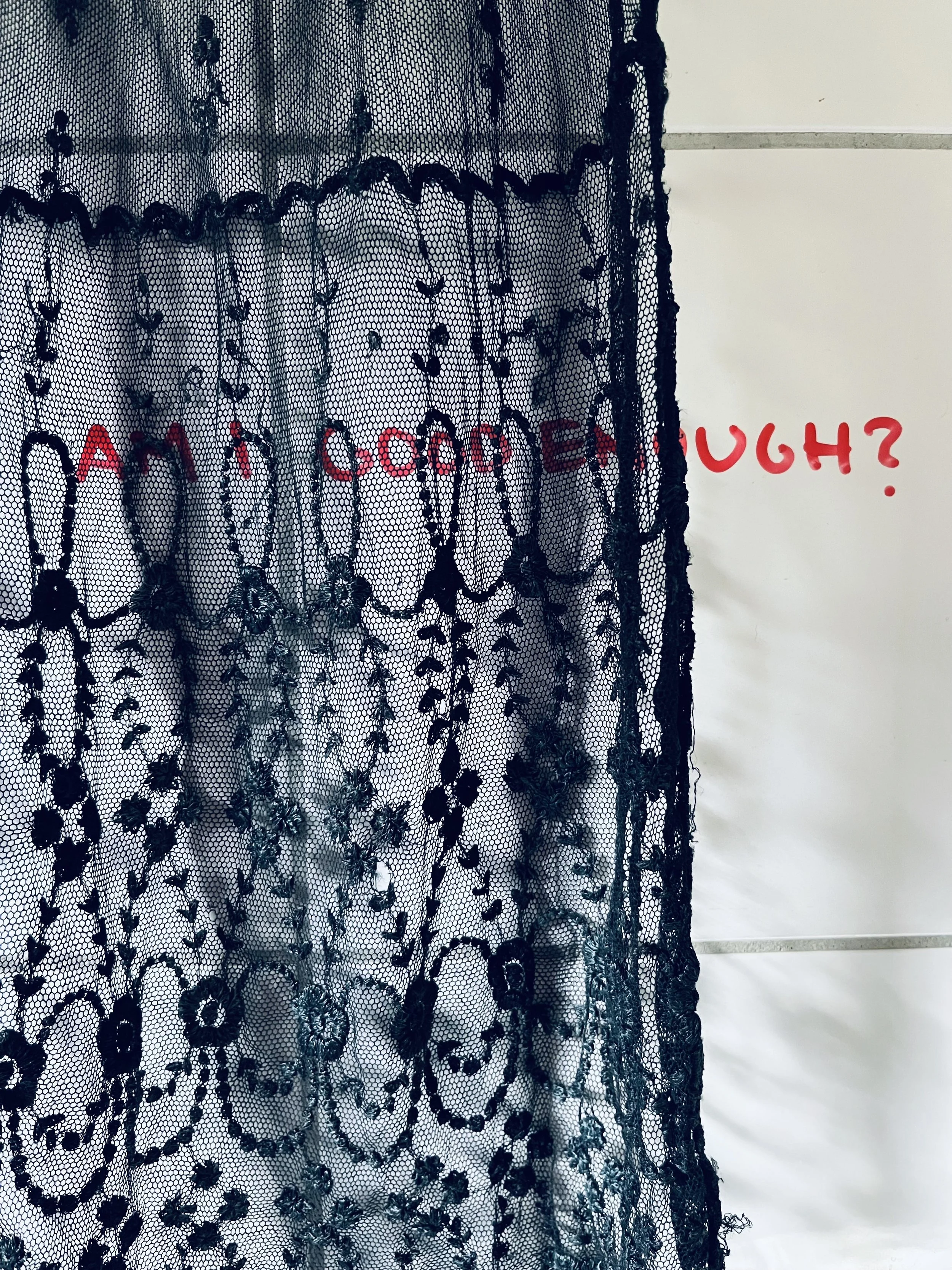 Black lace curtain with floral pattern hanging in front of a white wall with red handwritten text that reads 'AM I GOOD ENOUGH?'