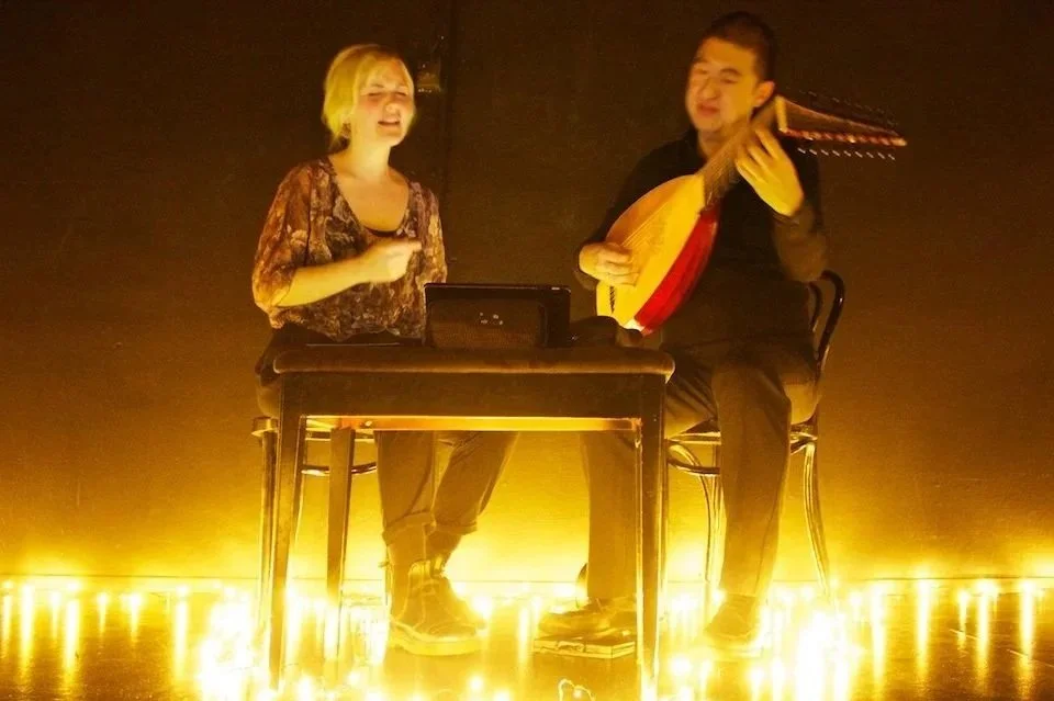 Two people performing music on a stage with warm yellow lighting. One person is singing and the other is playing a stringed instrument.