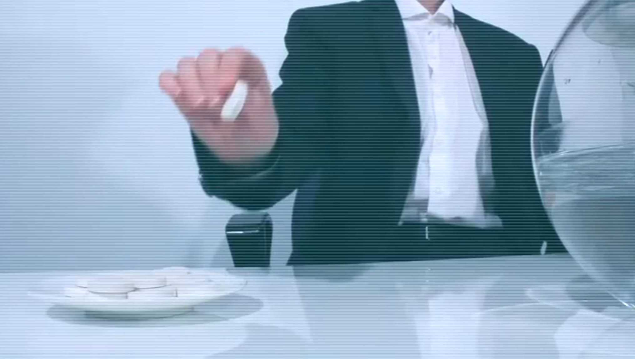 A person in a black suit and white shirt is sitting at a table with a stack of white round objects, possibly tablets, in front of them. The background is plain and light-colored.