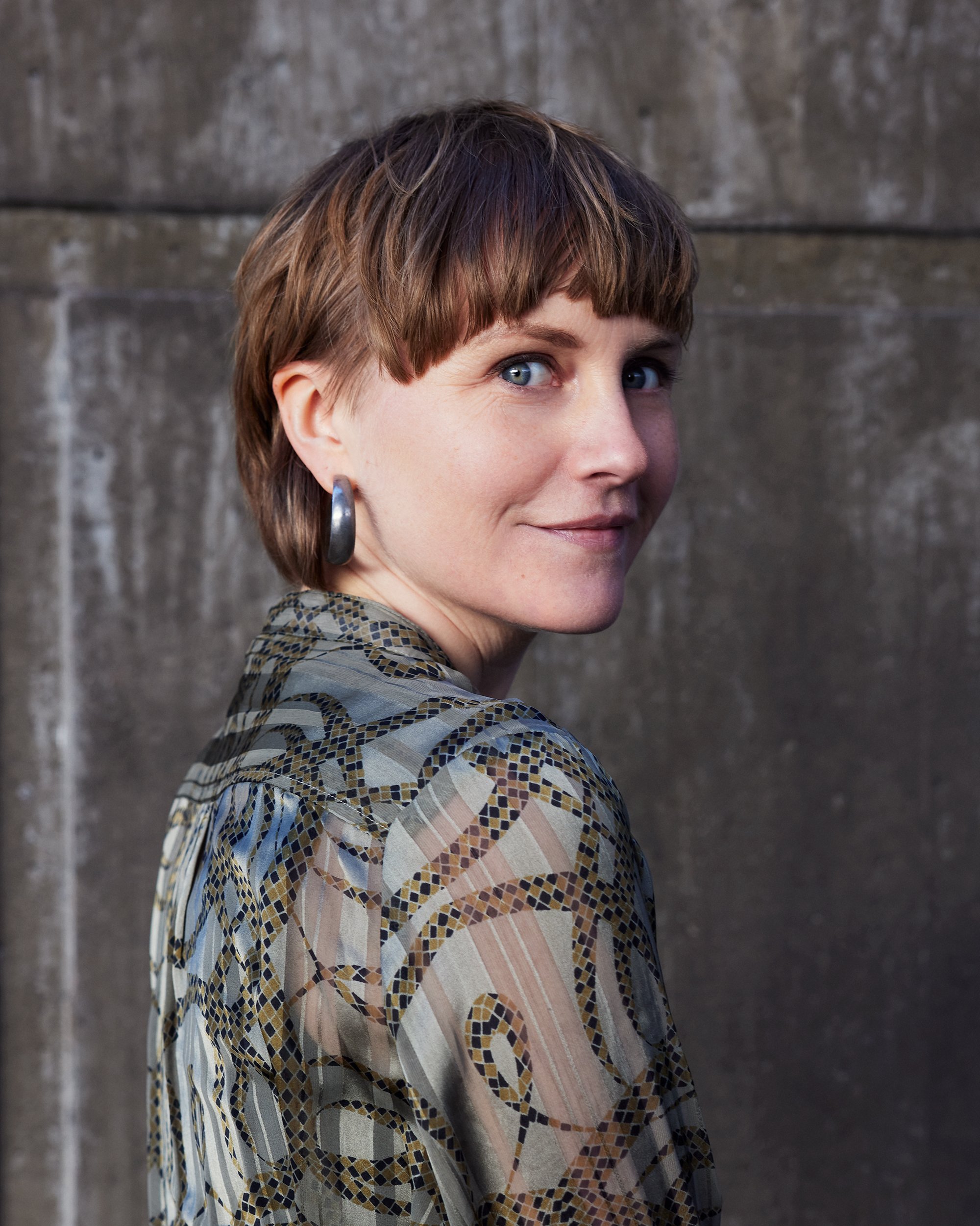 Close-up of a woman with short brown hair, wearing hoop earrings, smiling slightly, in front of a grey, textured stone wall.