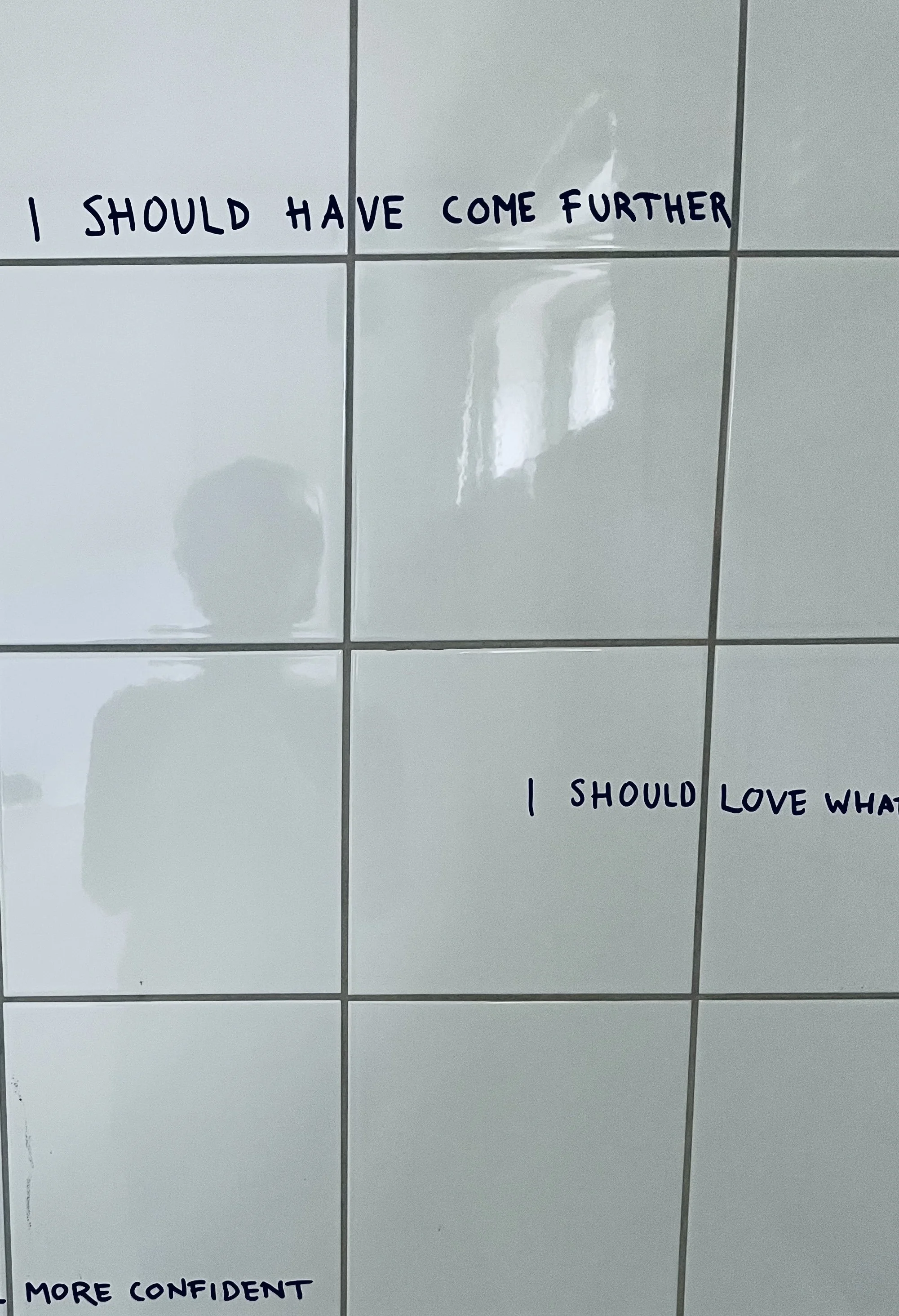 White tiled wall with handwritten statements including "I should have come further," "I should love what I do" and other words partially visible.