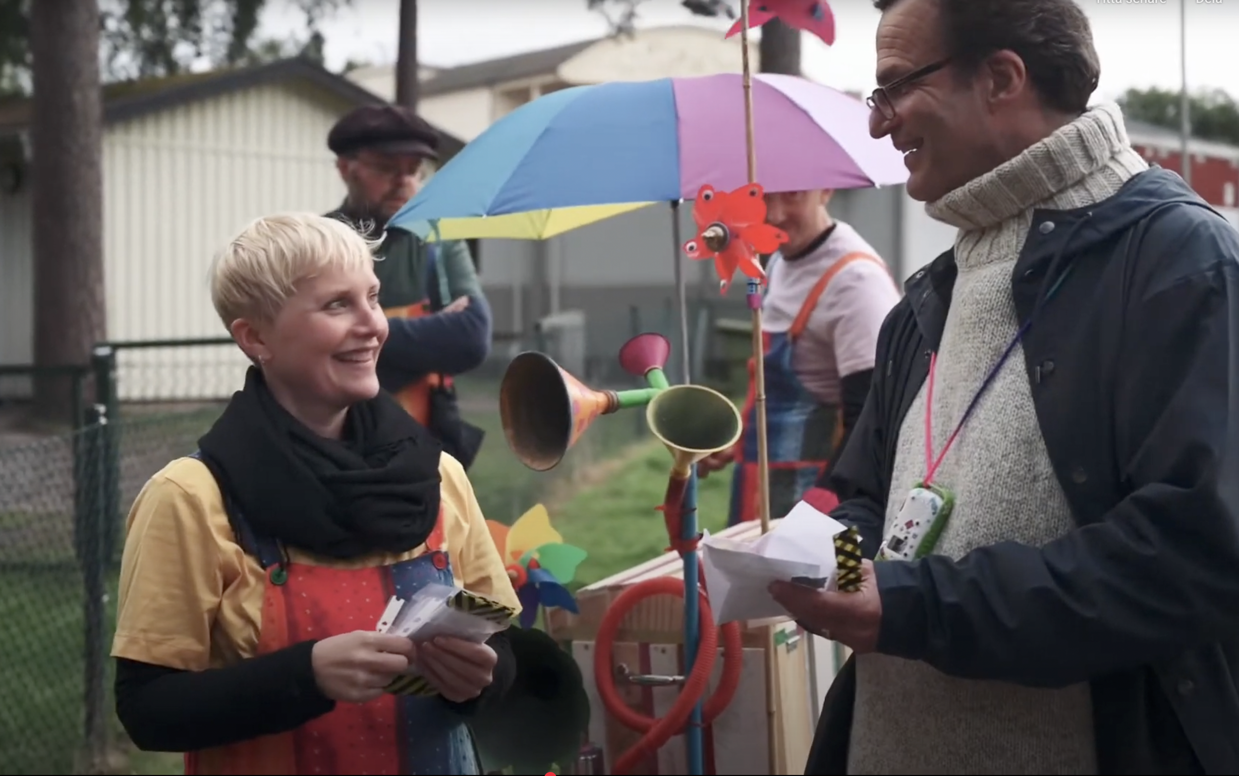 A woman and a man are smiling and talking outdoors. The woman is wearing a black scarf and a tan jacket, while the man has glasses, a knit turtleneck, and a dark jacket. 