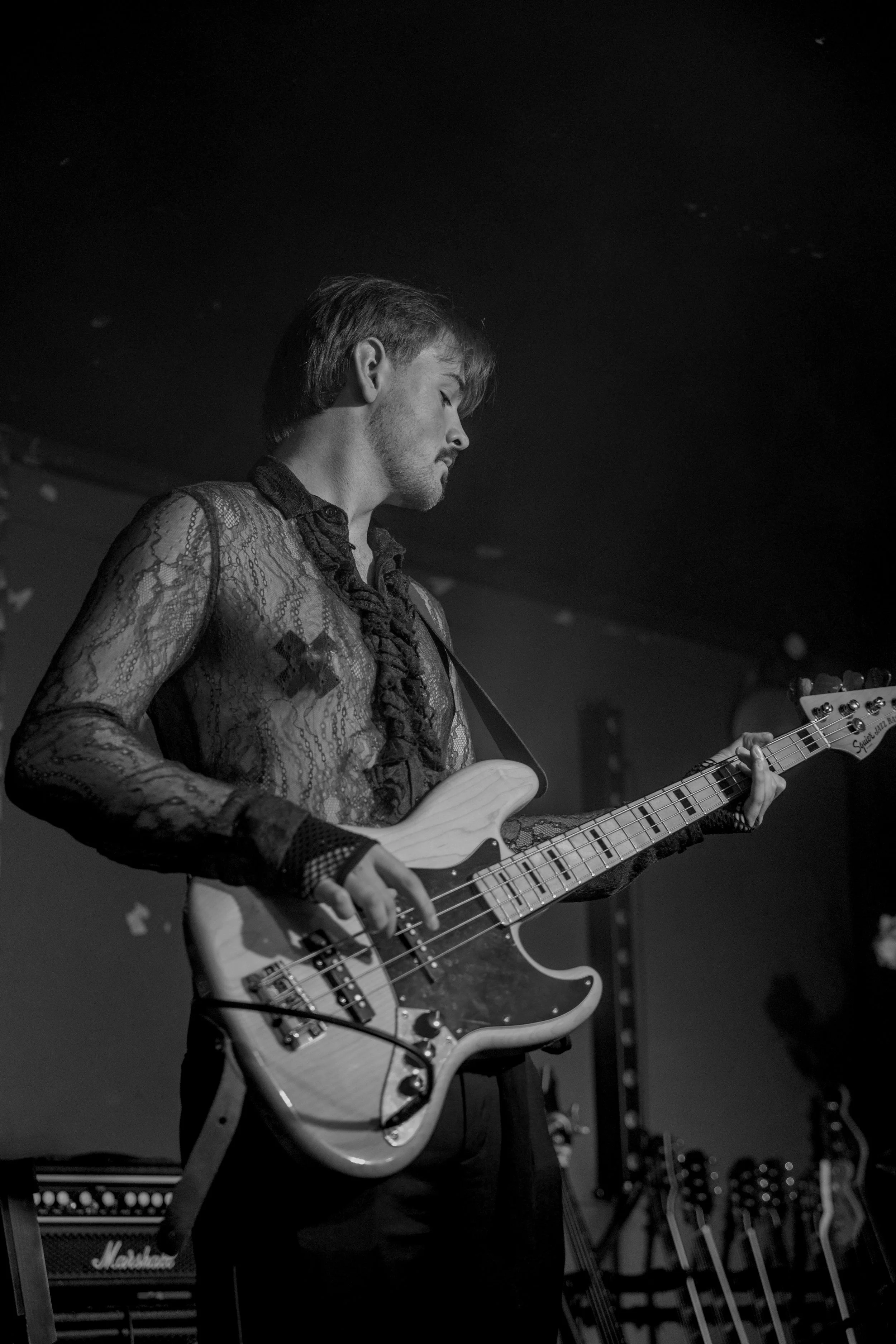 A black-and-white photo of a man playing an electric bass guitar on stage. He has short hair, a beard, and is wearing a lace or mesh shirt. In the background, there are musical equipment and amplifiers.