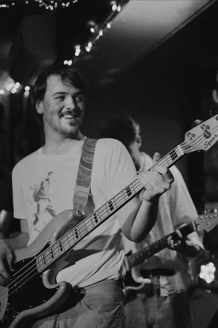 A man playing a bass guitar at a live music venue, smiling, with string lights overhead, in black and white.